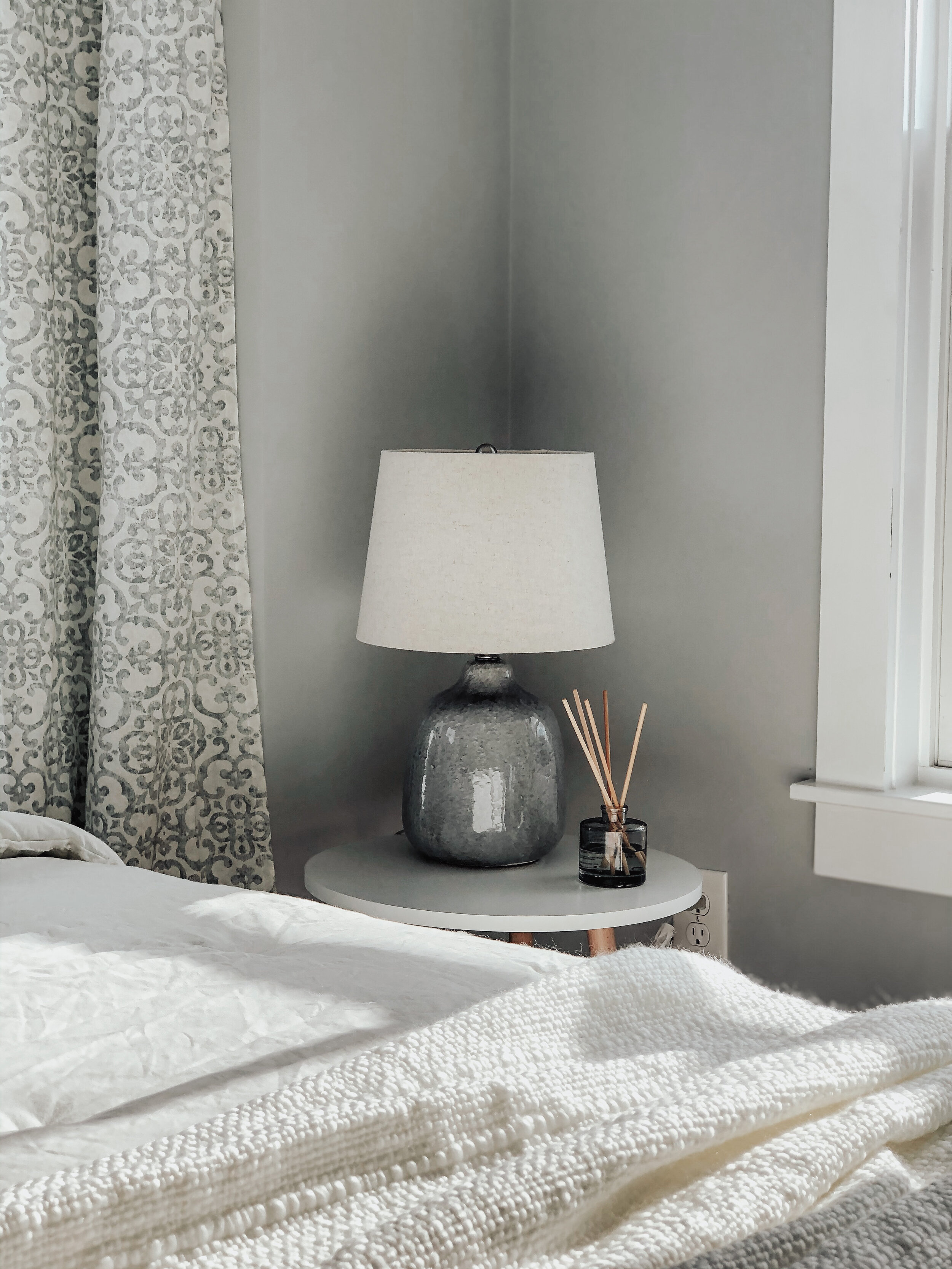 A cozy bedroom corner with a white bed, patterned curtains, a gray table lamp, and a small reed diffuser on a side table next to a window.