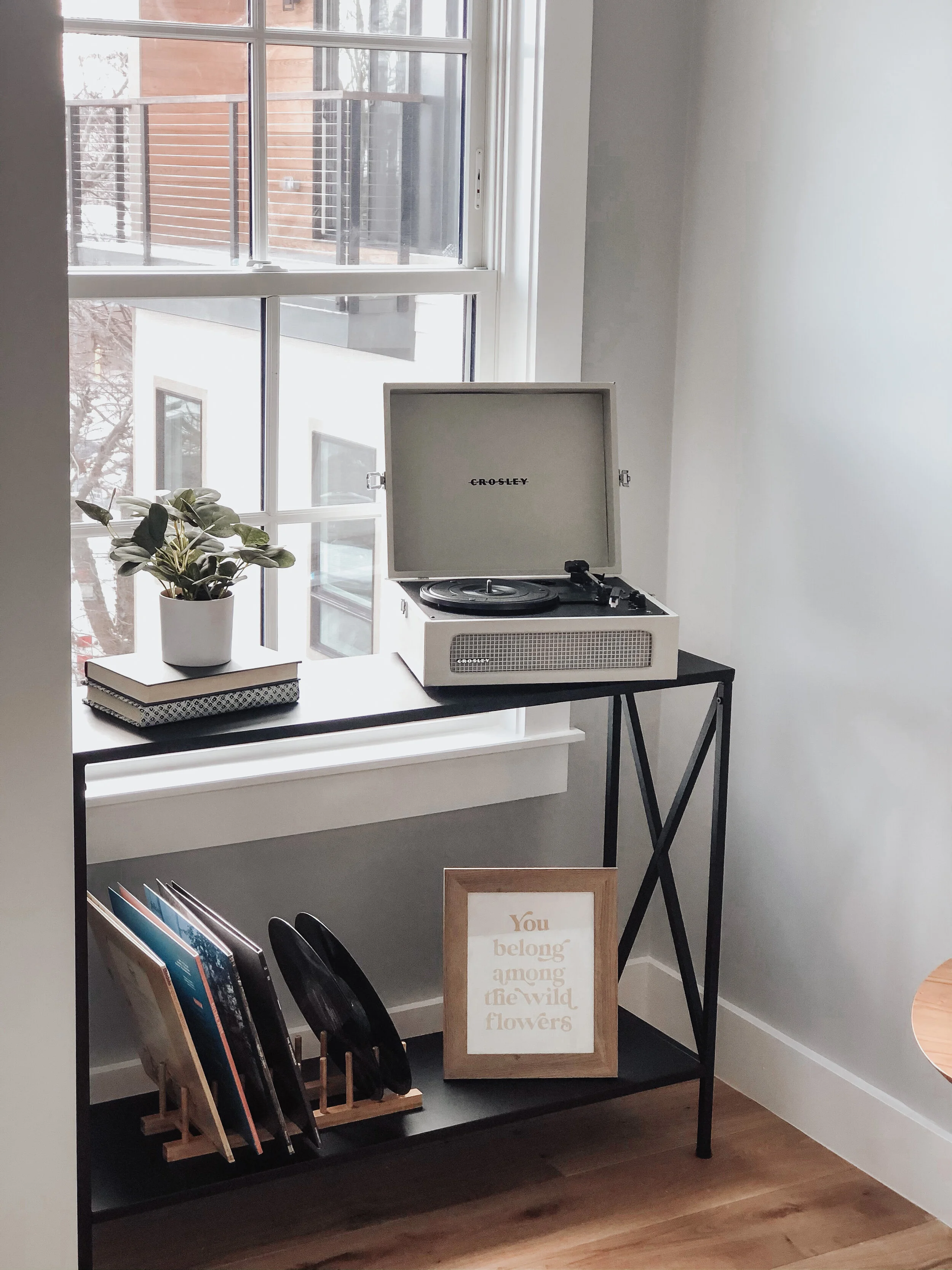 A vintage Crosley turntable on a black metal console table near a large window, with a potted plant, a stack of books, and a framed quote that reads, 'You belong among the wildflowers'.