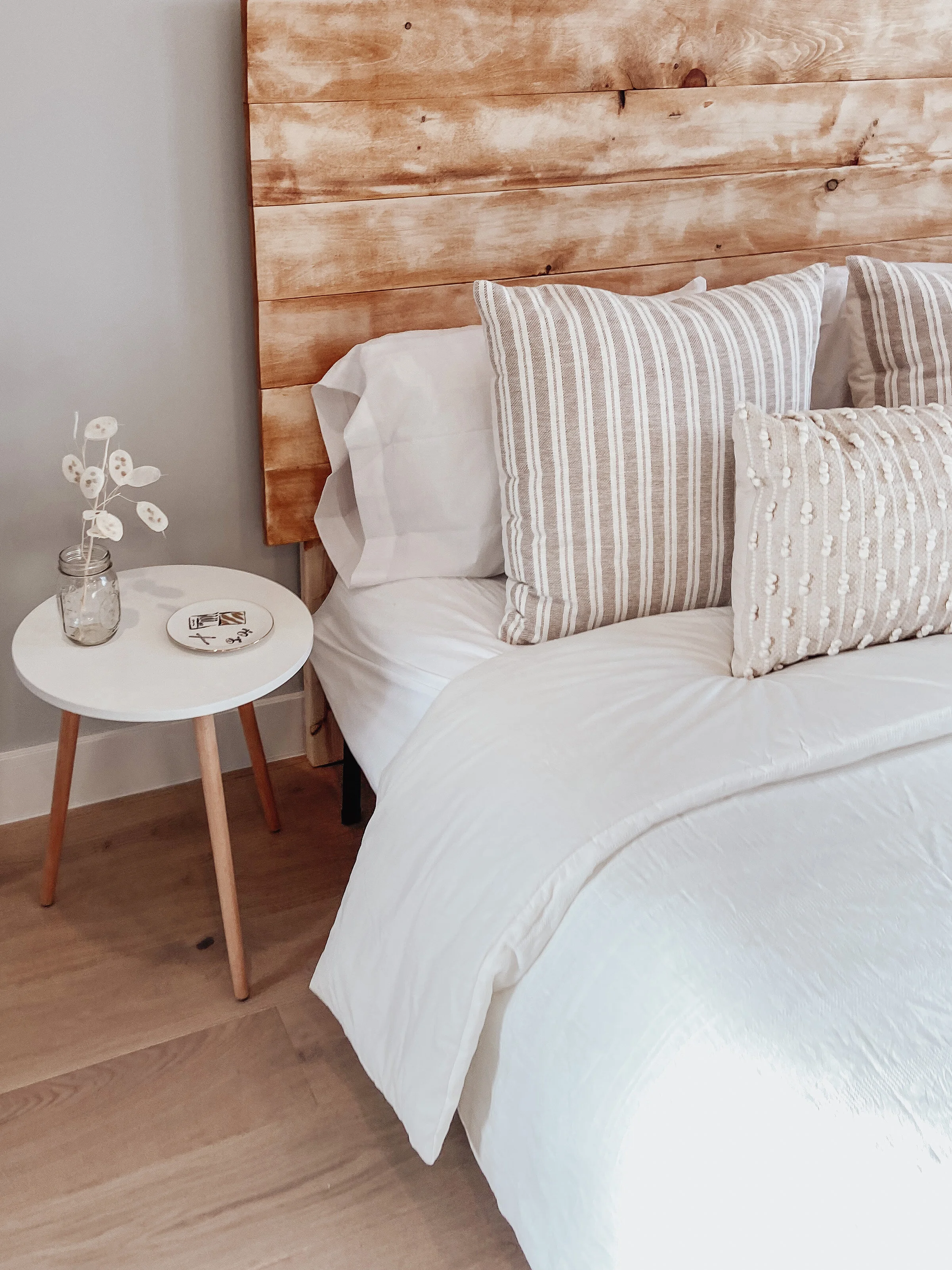 A cozy bedroom with a wooden headboard, white bedding, and decorative pillows. A white side table with a small vase and a decorative plate is next to the bed.