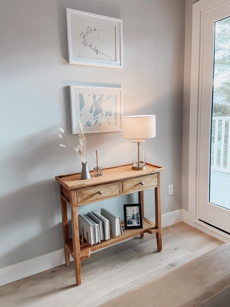A wooden console table with two drawers and a lower shelf, holding a vase with dried grass, a small reed diffuser, and a modern table lamp. Above the table are two abstract art pieces, and there is a framed photograph on the lower shelf. To the right