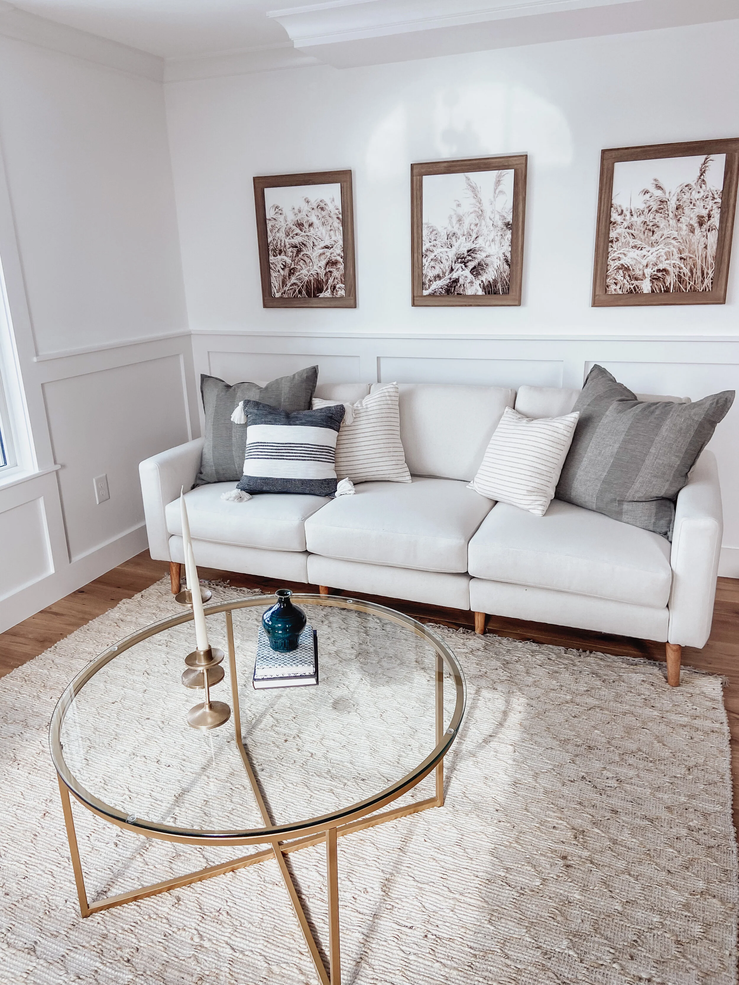 Living room with a white sofa, striped and plain pillows, framed artwork of grass or plants on the wall, gold coffee table with candles and a book, textured beige rug, natural light from a window.
