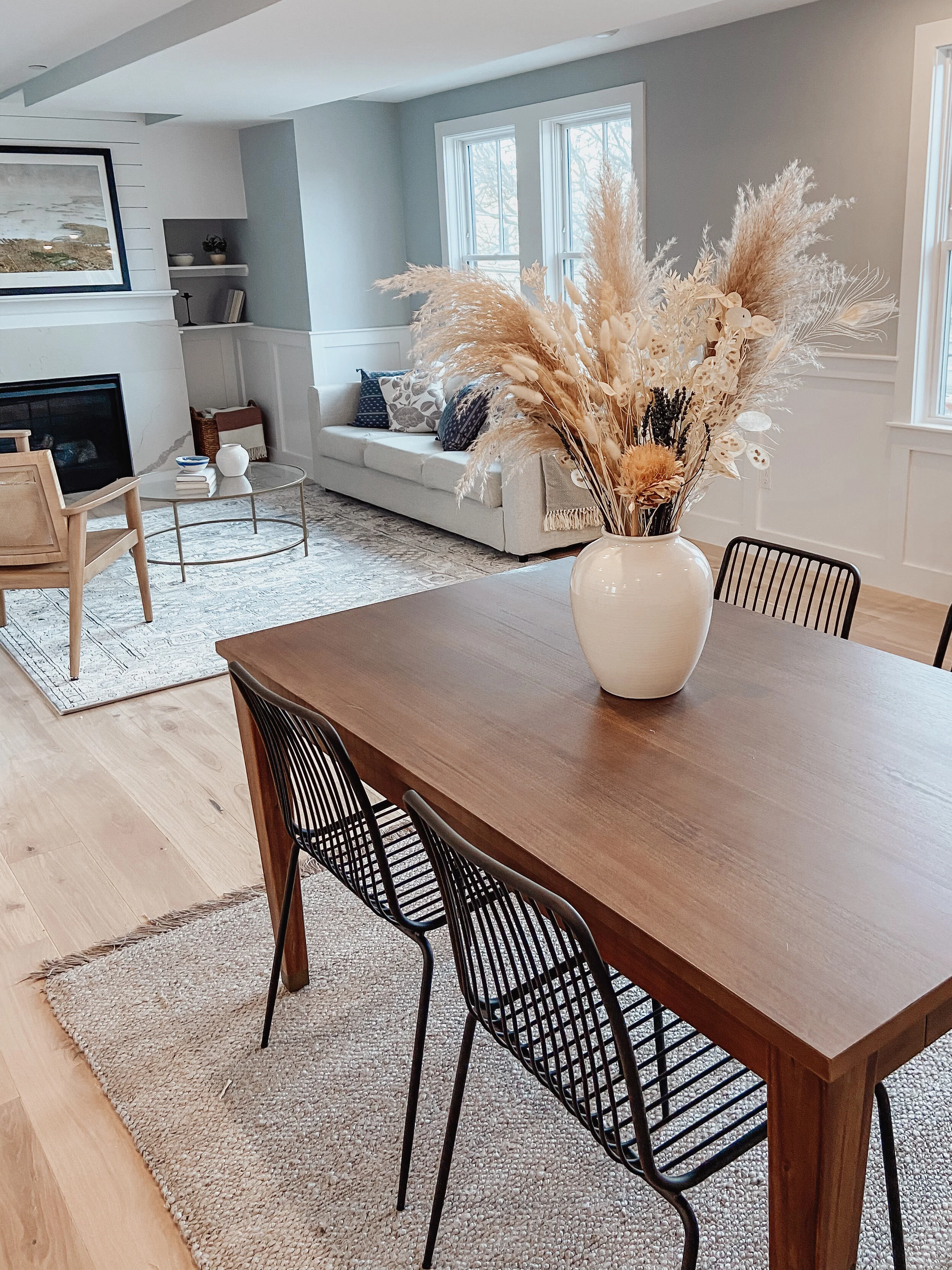 Living room with wooden dining table, black wire chairs, large white vase with dried pampas grass, sofa with decorative pillows, fireplace, window with winter scene outside, and neutral decor.