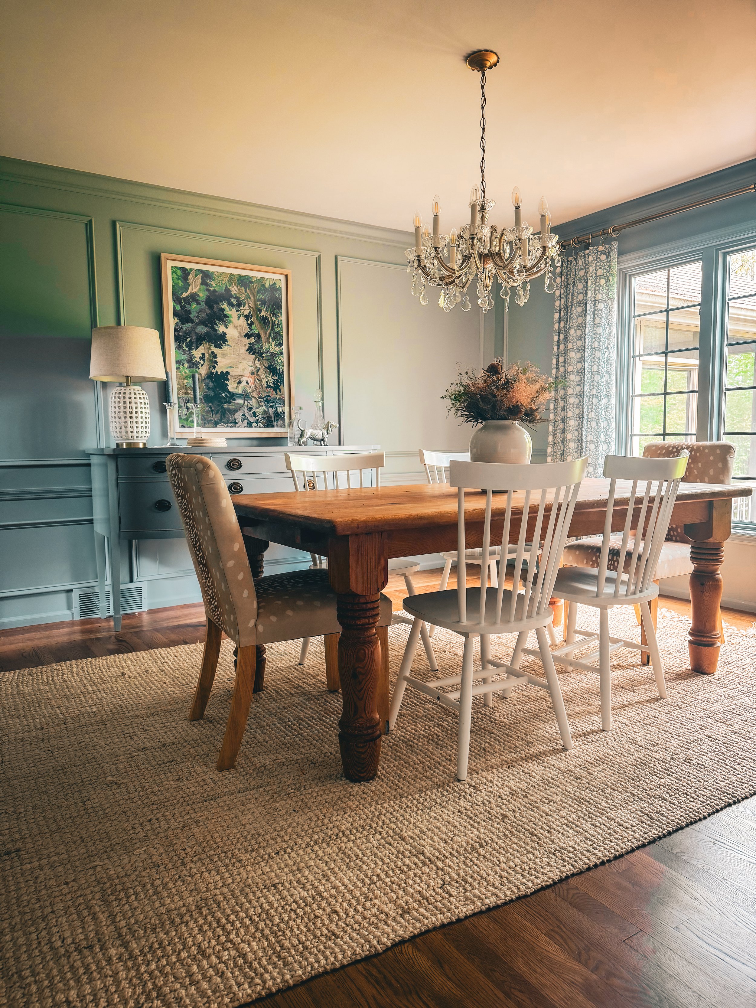 A dining room with a wooden table, six chairs, a chandelier, a sideboard with a lamp, a framed picture, a large vase with dried flowers, and a window with curtains, sunlight coming through.