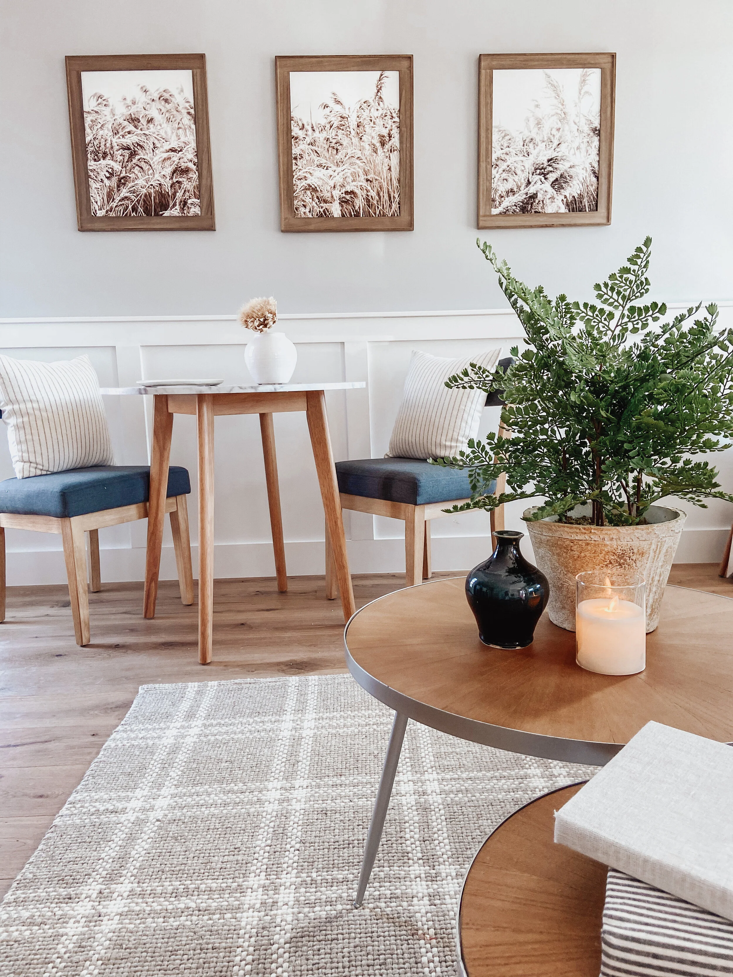 A cozy living room corner with a small white table, two chairs with blue cushions, pale striped pillows, a large green plant in a pot, a black vase, a lit candle, and three framed pictures of tall grasses on the wall.
