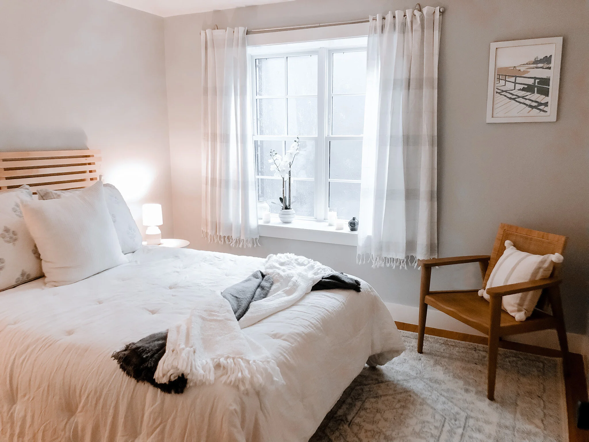 Bright bedroom with a bed, white bedding, and pillows. A side table with a lamp, and a wooden chair with a cushion. A window with white curtains, a potted flower, and candles on the windowsill. Wall frame art and light-colored walls.