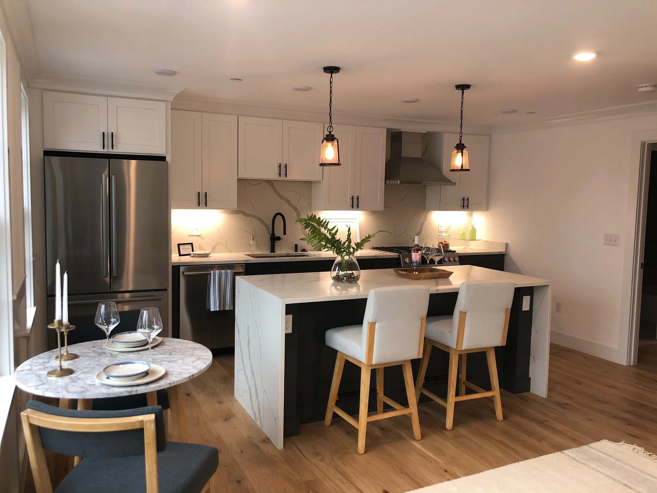 Modern kitchen with white cabinets, marble backsplash, stainless steel refrigerator, black accents, island with barstools, small dining table with chairs, and hardwood floors.