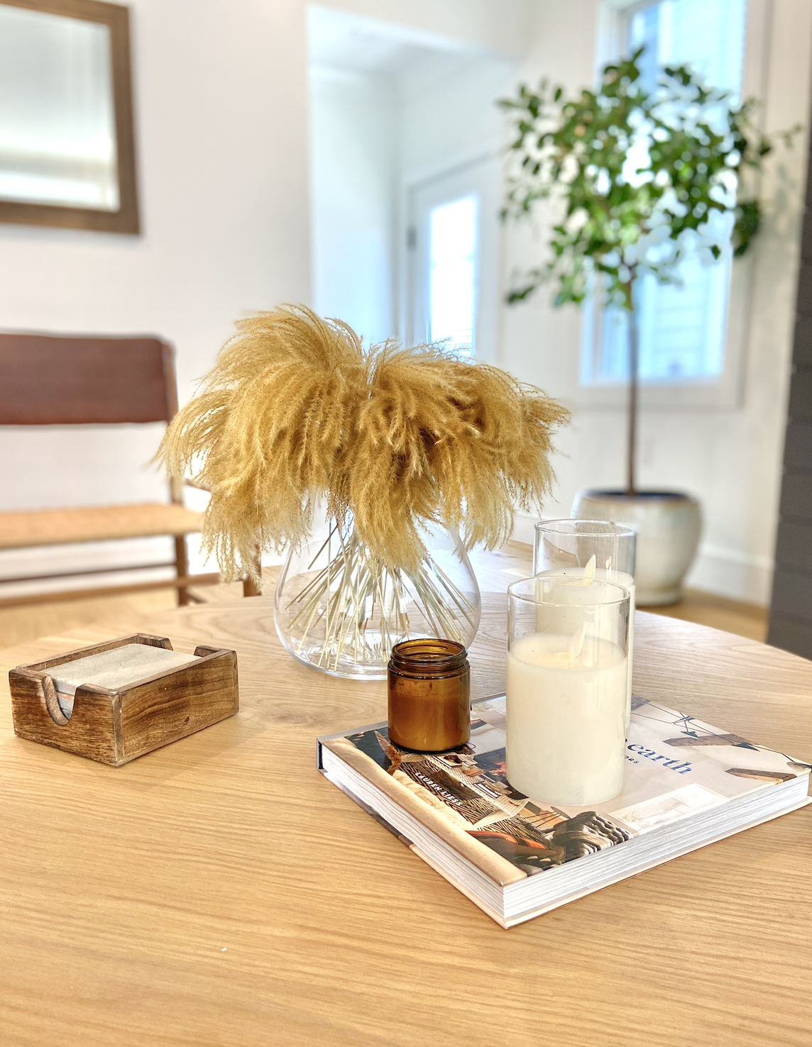 A wooden table with a magazine, two white candles in glass holders, a small amber glass jar, a round glass vase with yellow dried flowers, a wooden tissue box, and a potted plant in the background.