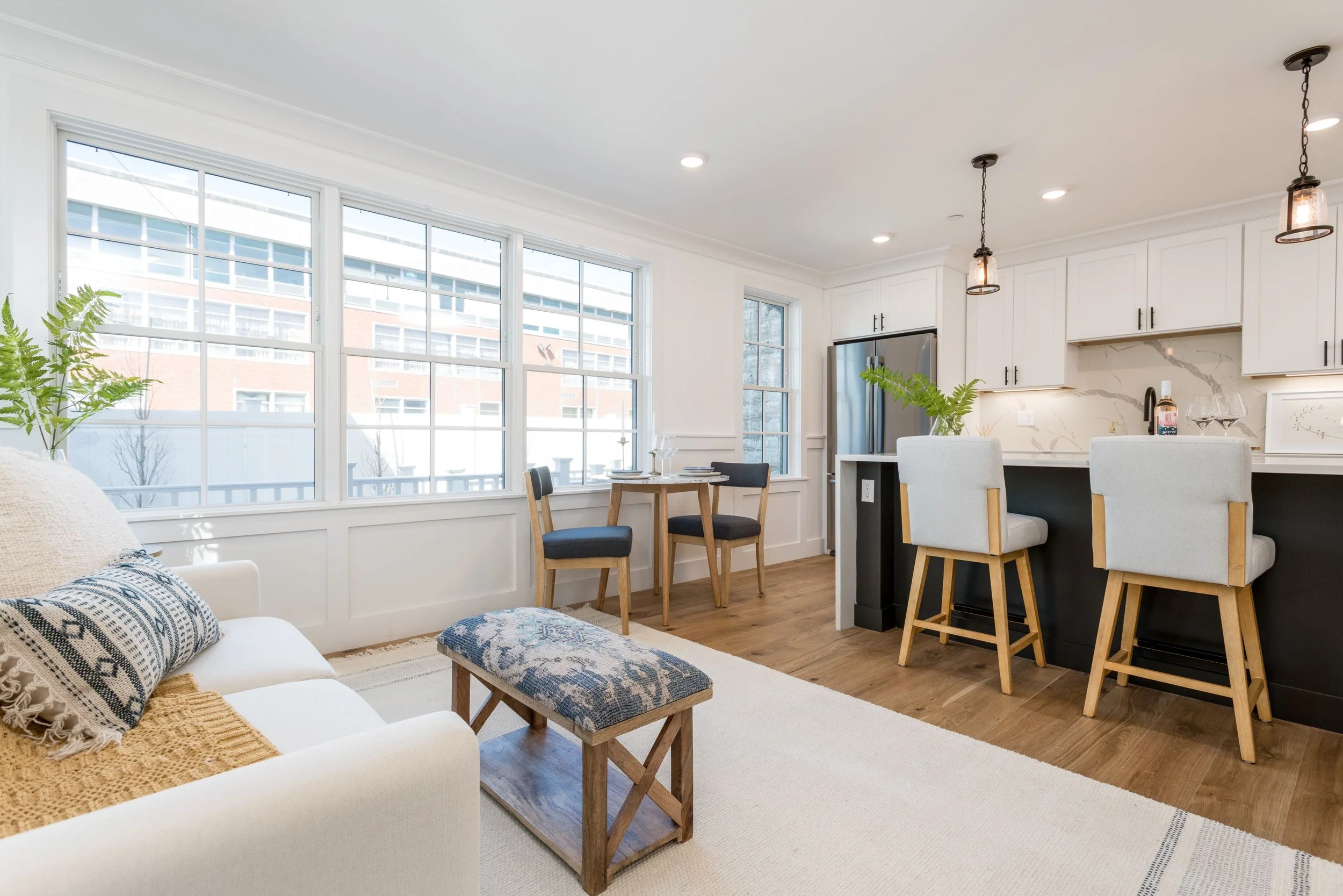 Open-concept living room and kitchen with large windows, white cabinetry, island with bar stools, small dining table, and wooden flooring.