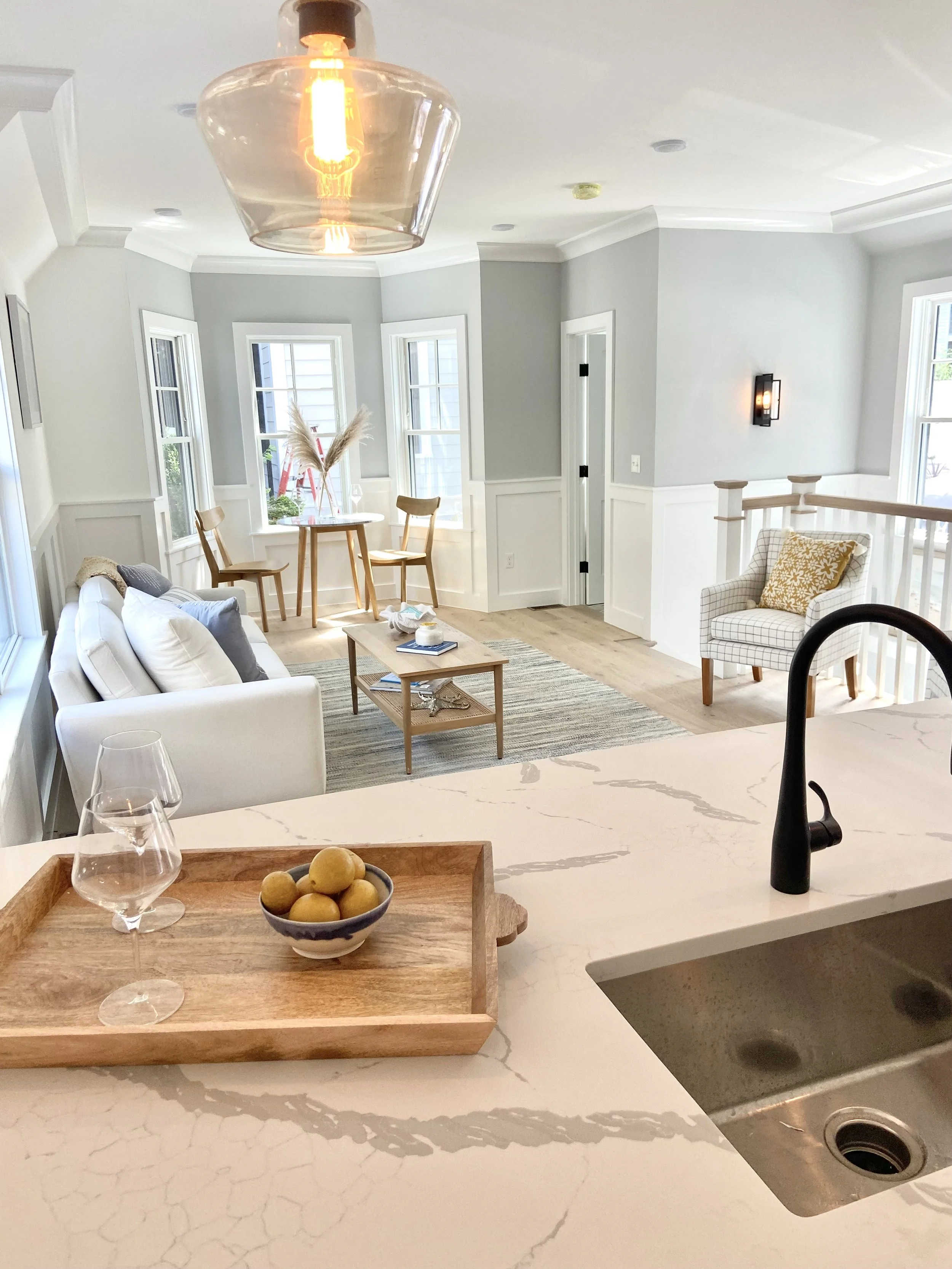 Open-concept living room and kitchen with natural light, white walls, light wood flooring, and modern fixtures, viewed from a kitchen island with a bowl of lemons and glasses on a wooden tray in the foreground.