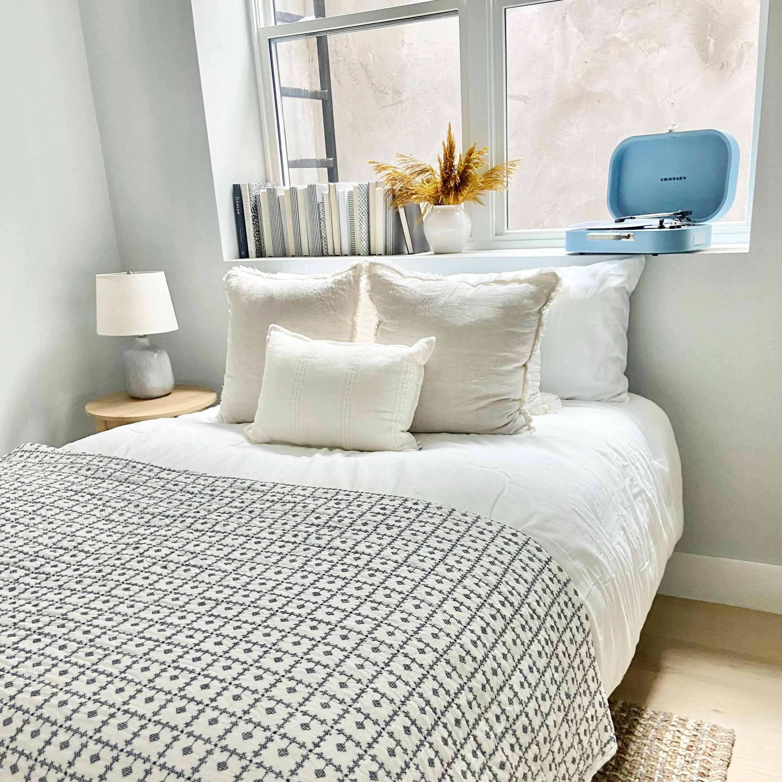 A cozy bedroom with a white bed, decorated with cream pillows and a black and white patterned blanket. A small wooden side table with a gray lamp is next to the bed. A window behind the bed has a vase with dried flowers, a row of books, and a blue re