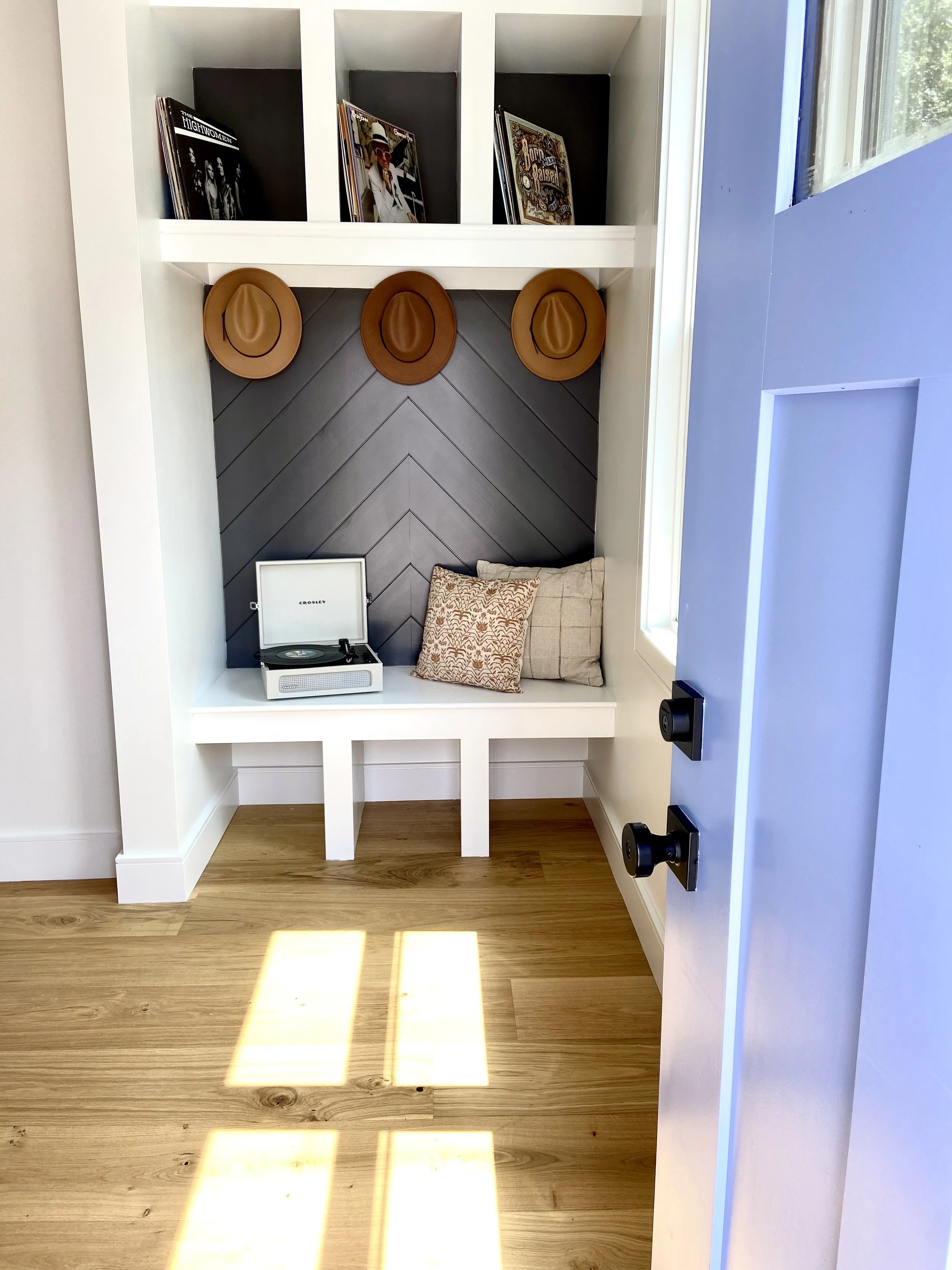 A small cozy nook with a built-in bench, black shiplap wall, pillows, a vintage record player, and three hats hanging on the wall. There are shelves above with vinyl records and magazines, and sunlight streaming through a nearby window.