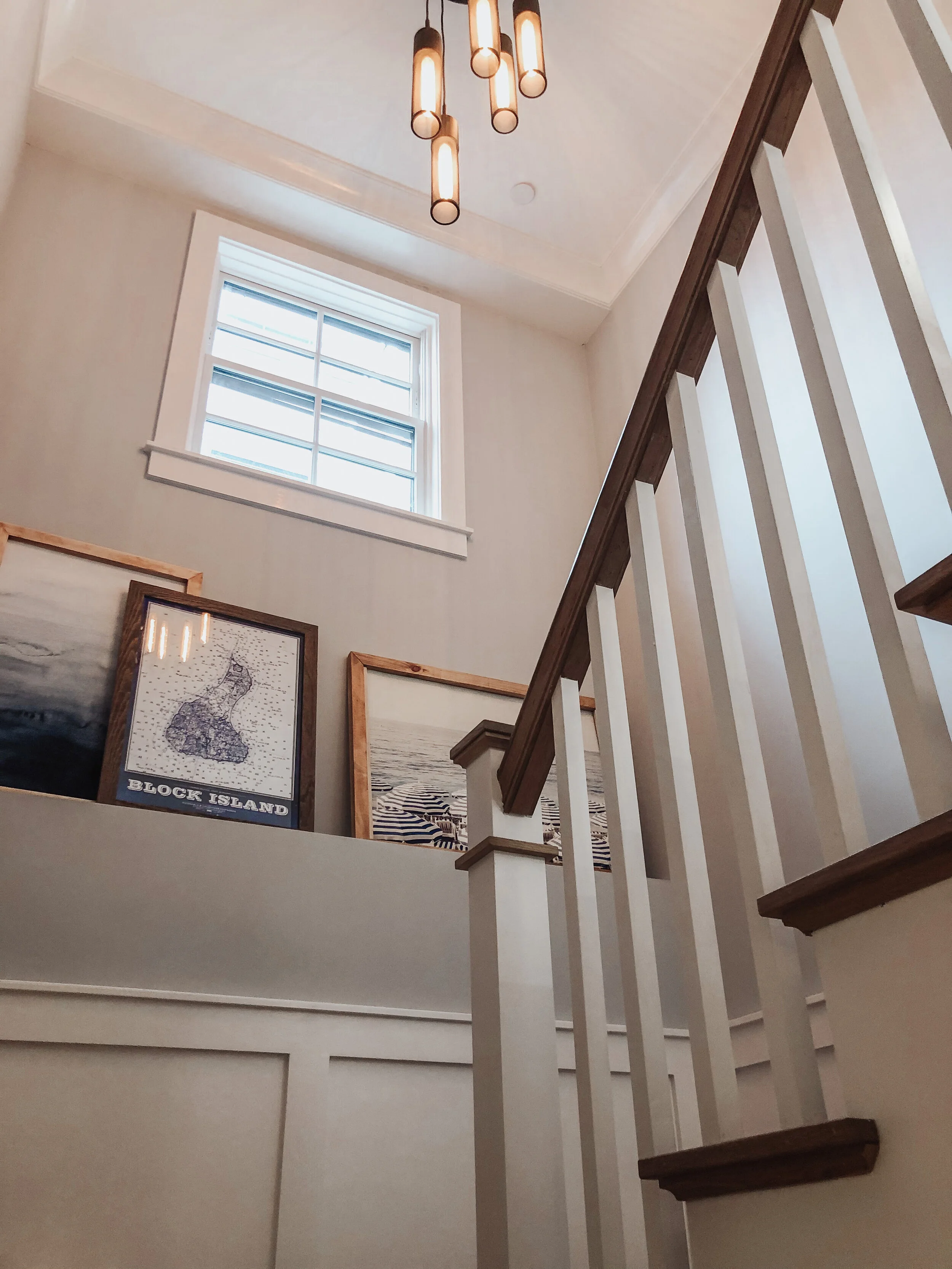 Interior view of a staircase landing with framed artwork and a window, featuring a modern hanging ceiling light, wooden chair rail, and white walls.