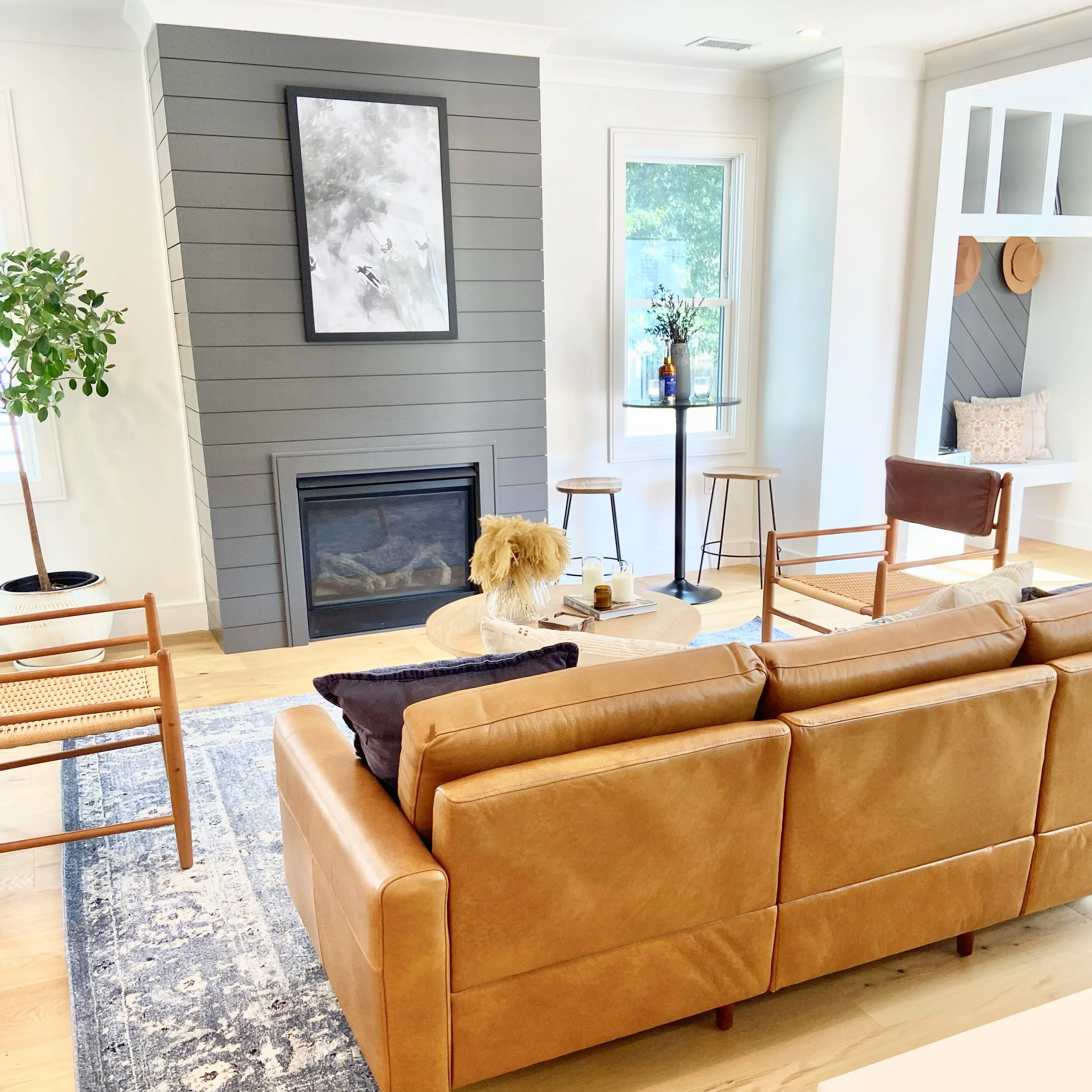 A cozy living room with a tan leather sofa, a round coffee table, a gray shiplap fireplace with a black framed photo above, a potted plant on the left, and a window with a small tall table and stools in the background.