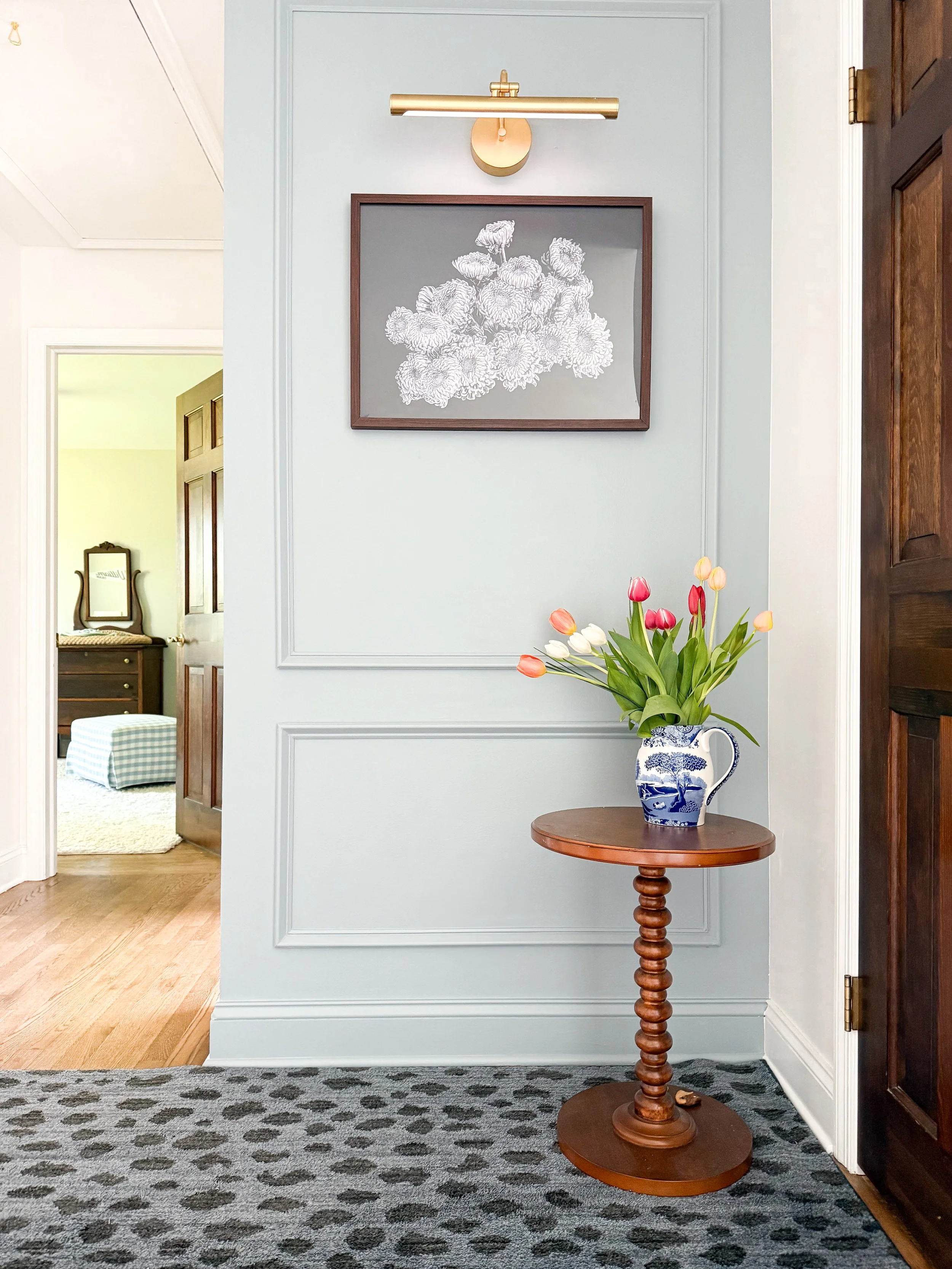 A small round wooden table with a blue and white porcelain pitcher filled with pink and white tulips, situated against a pastel green wall with decorative molding. A framed botanical print hangs above, illuminated by a gold picture light. Part of an 