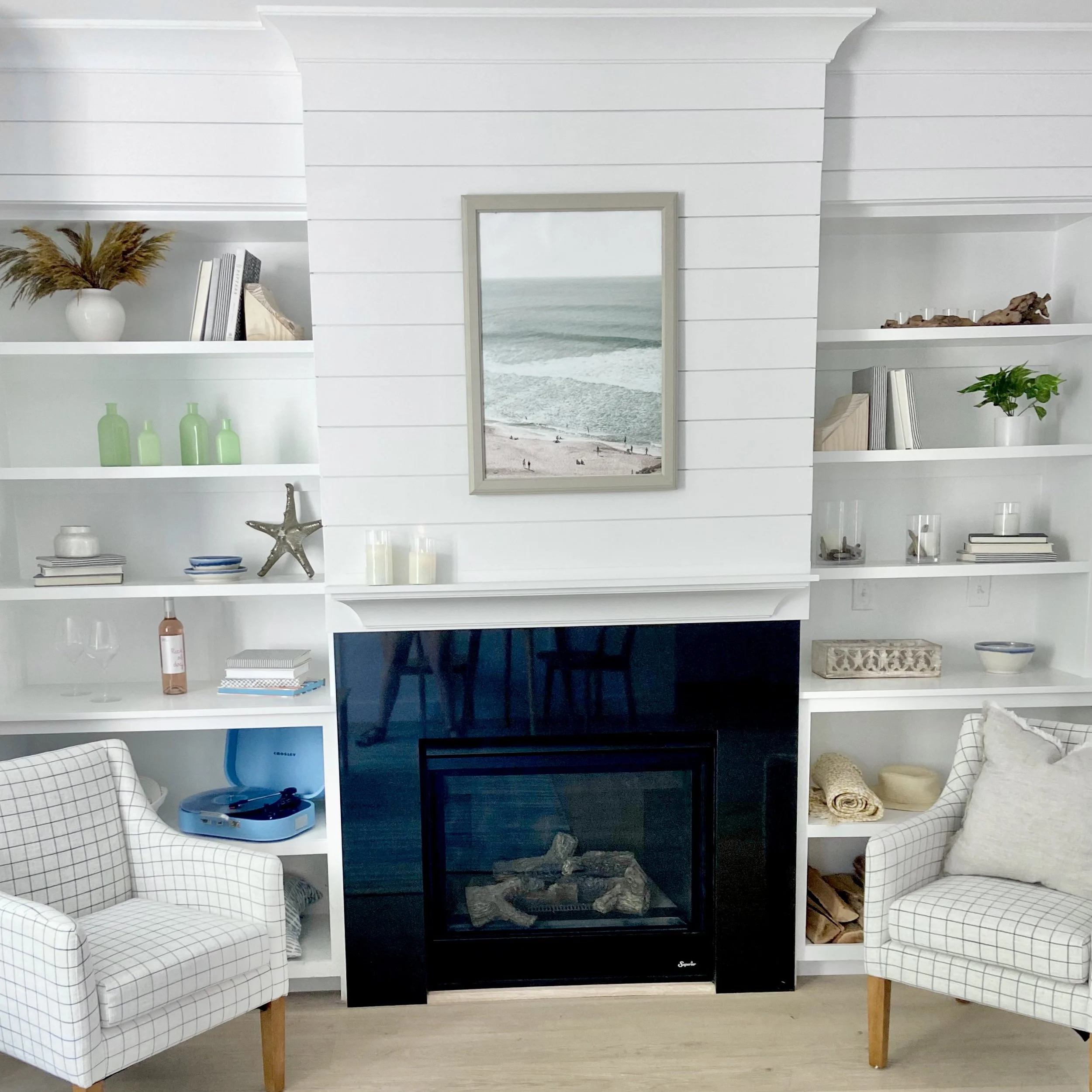 Living room with a white built-in bookshelf surrounding a black fireplace, a framed beach scene picture above the mantel, two plaid armchairs with pillows, and decorative items including books, bottles, a starfish, and candles.