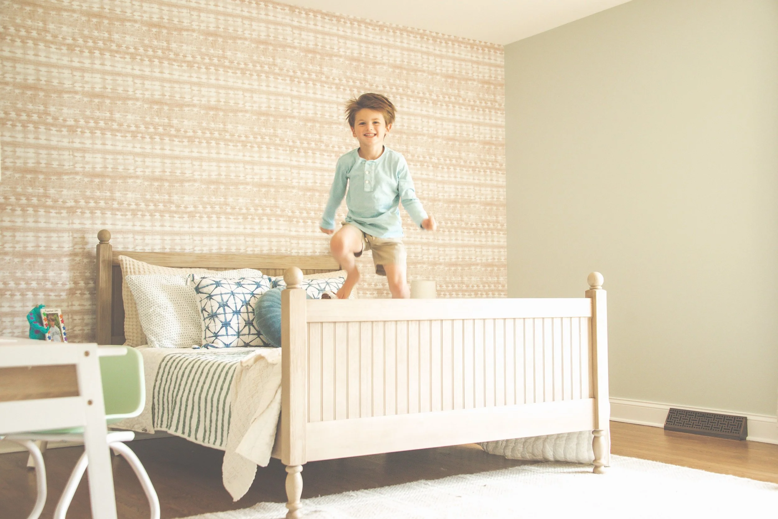 A young boy jumping on a bed in a softly decorated bedroom with patterned wallpaper and a light-colored wall.