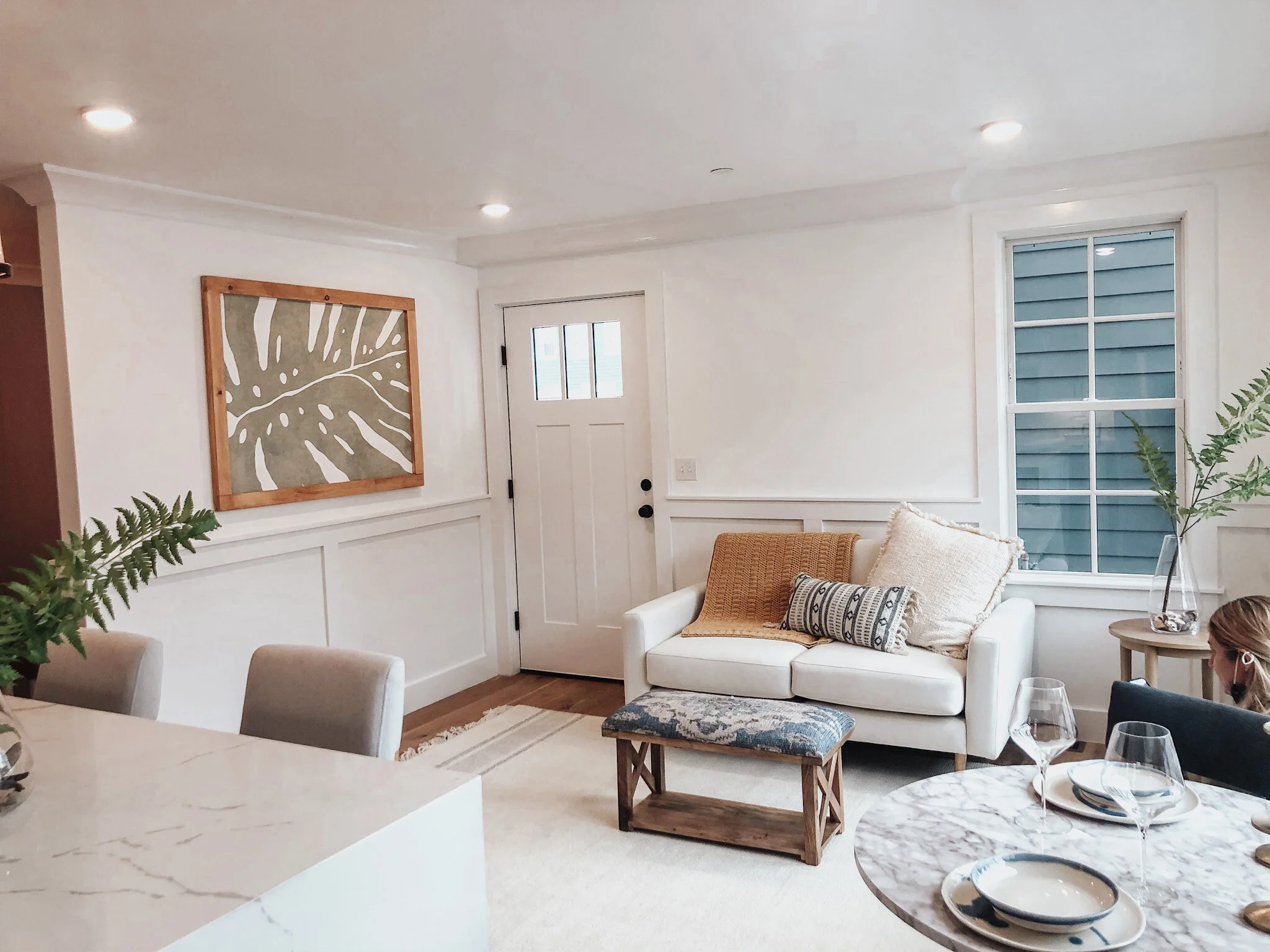 Bright living room with white walls, large window, beige sofa with throw pillows, wooden door, wall art, marble-topped dining table with glassware, and a person partially visible