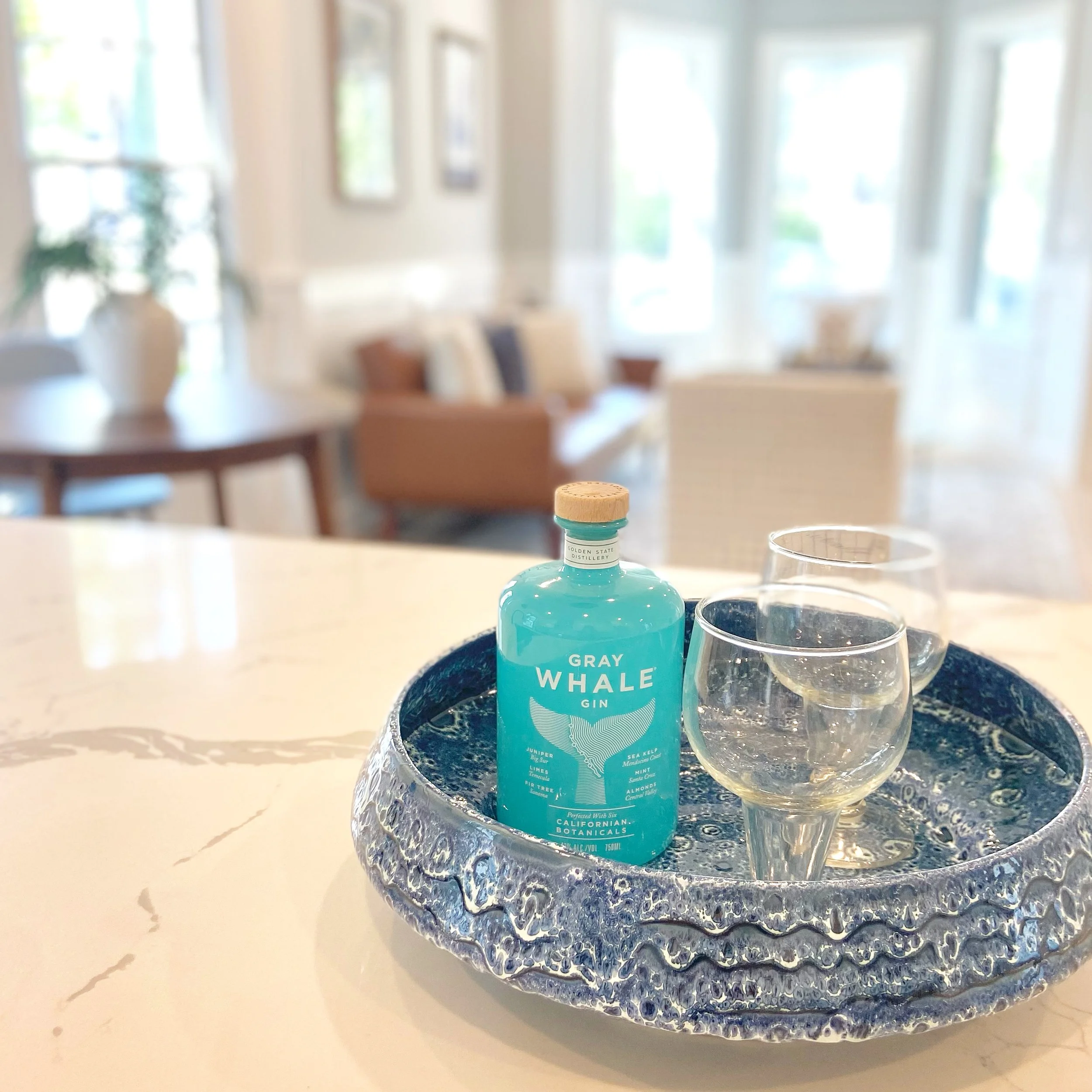 A bottle of Gray Whale Gin and two clear glasses filled with water on a decorative blue tray placed on a white countertop in a bright, modern living room.