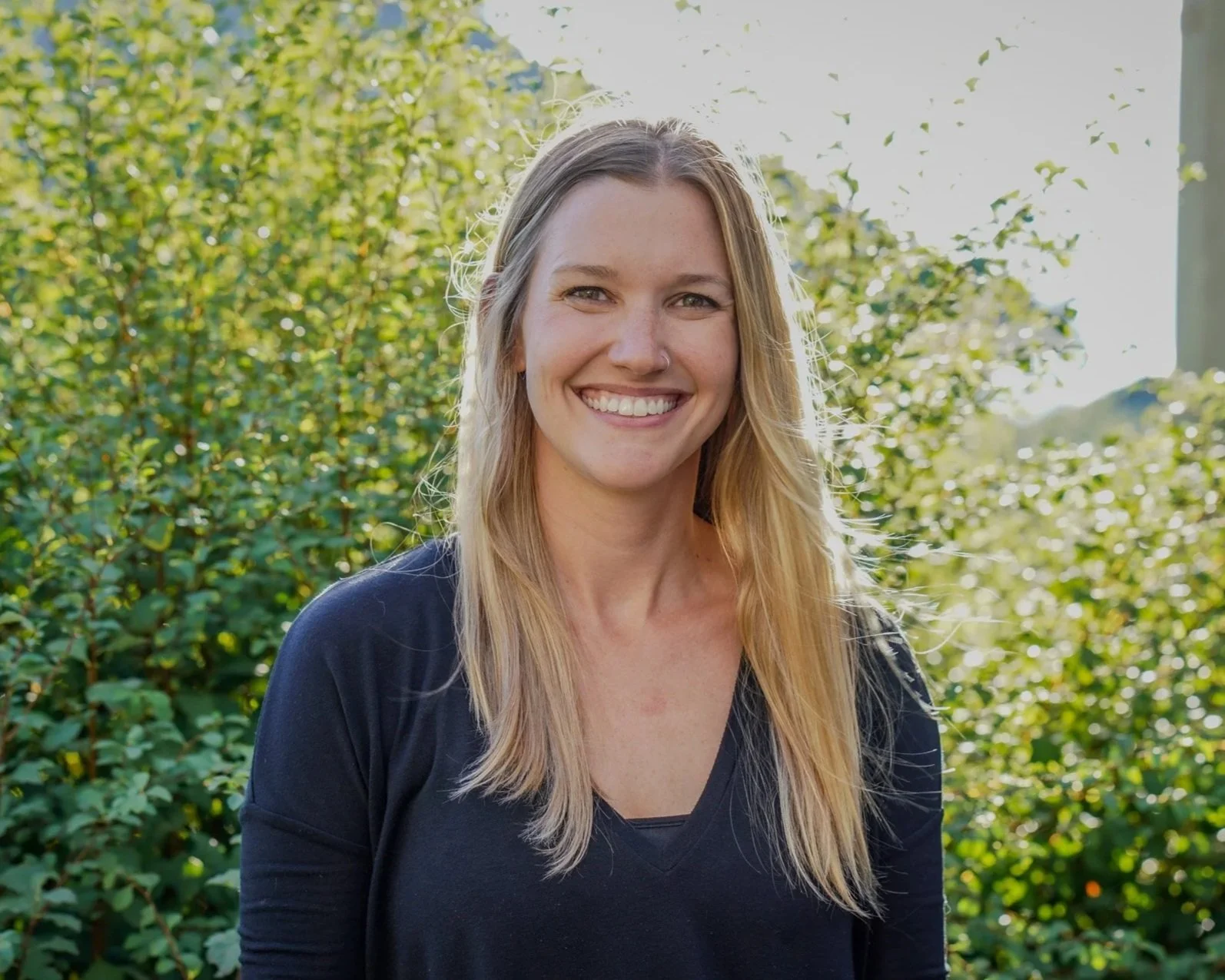 A young woman with long blonde hair and a bright smile standing outdoors with green bushes and sunlight in the background.