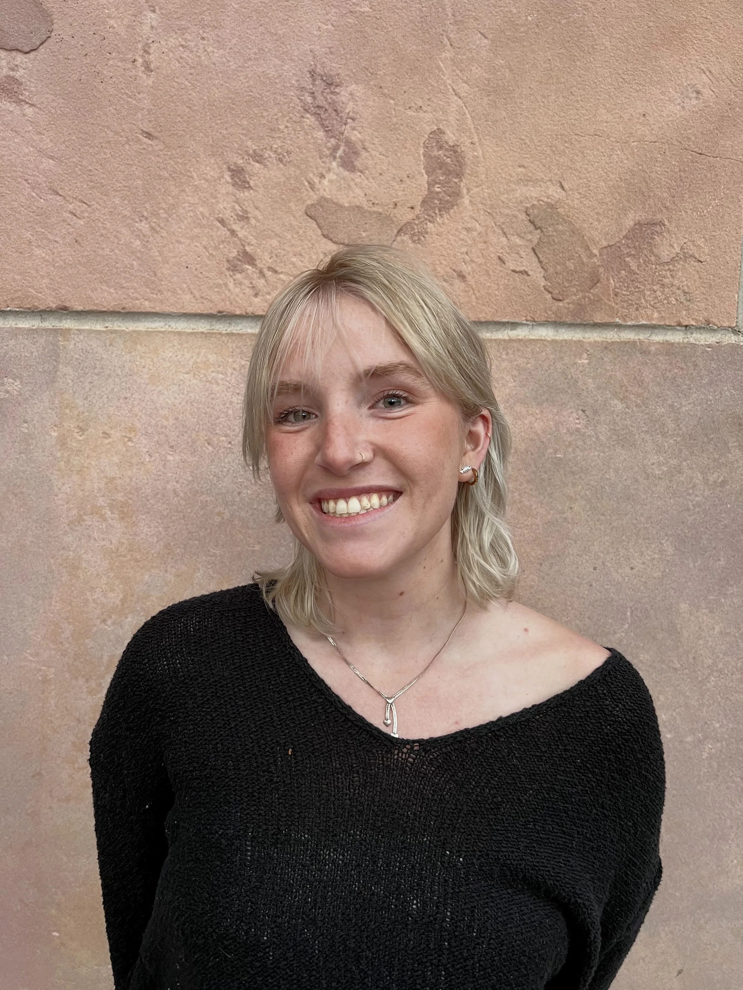 A smiling young woman with blonde hair, wearing a black knit top, silver jewelry, and earrings, standing against a textured pinkish-beige stone wall.
