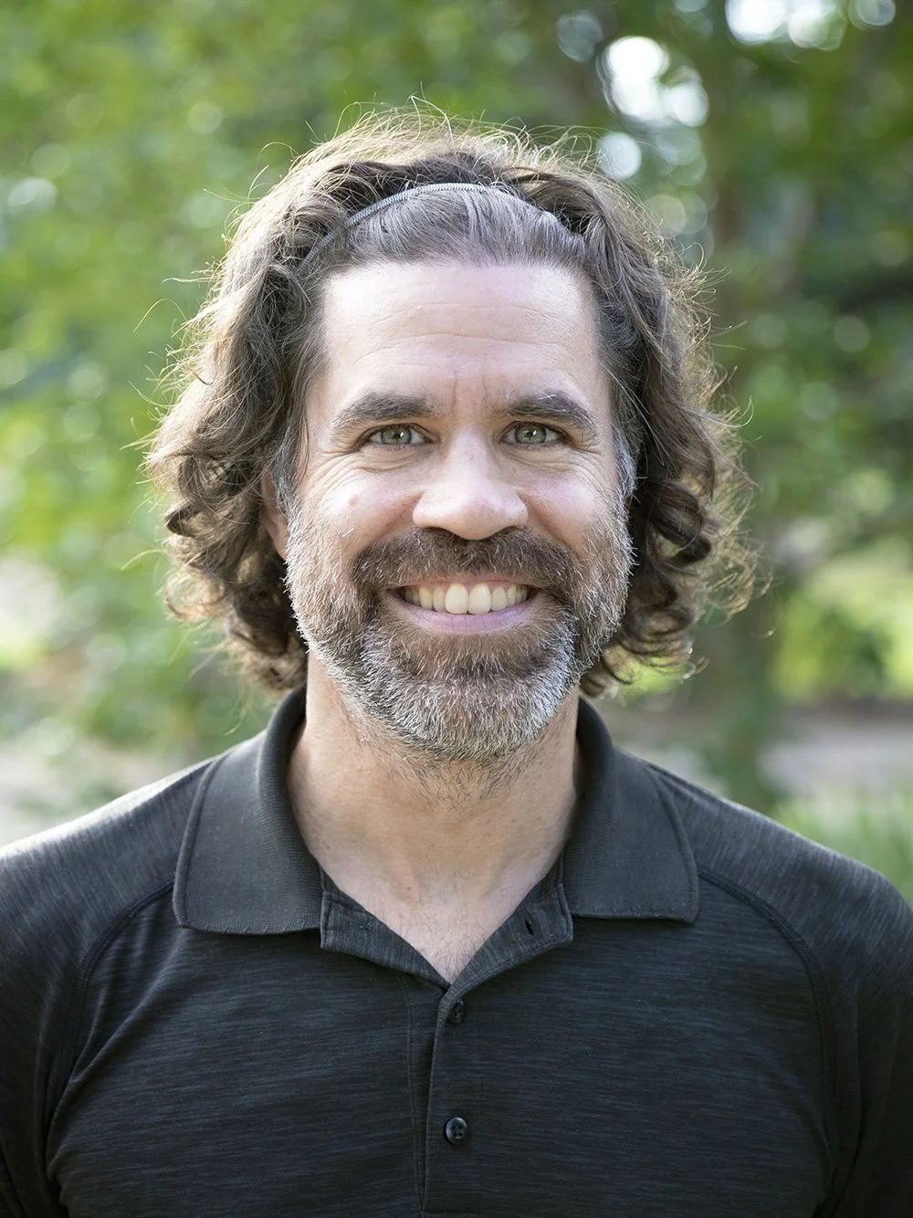 A smiling man with wavy, shoulder-length brown hair, a beard, and green eyes, wearing a black polo shirt, outdoors with green foliage in the background.
