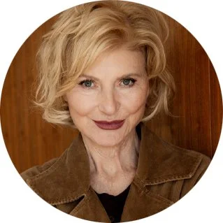 A woman with short blonde hair and dark lipstick smiling at the camera, wearing a brown jacket against a wooden background.