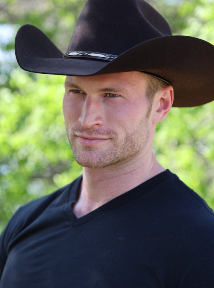 A man wearing a large black cowboy hat and a black V-neck shirt outdoors with blurred green foliage in the background.
