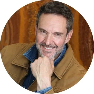 Portrait of a smiling middle-aged man with gray hair and beard, wearing a tan jacket and blue shirt against a warm brown background.