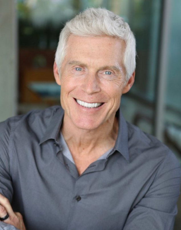 A smiling older man with gray hair wearing a gray button-up shirt in a bright indoor setting.