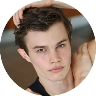 Close-up portrait of a young man with short dark hair, looking into the camera with a serious expression, resting his hand on his head.