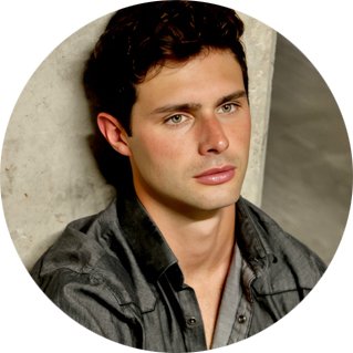 A young man with dark hair and fair skin, wearing a dark shirt, sitting against a light-colored wall.