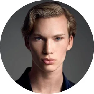 Close-up of a young man with blond hair and blue eyes, looking directly at the camera against a dark gray background.