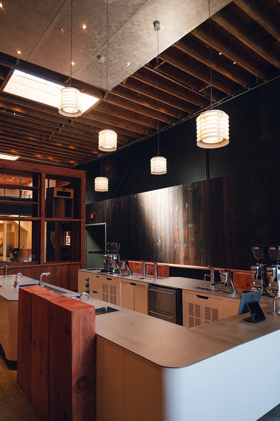 Interior of a modern coffee shop or cafe with wooden accents, pendant lights hanging from a ceiling with exposed beams, and a counter with espresso machines.