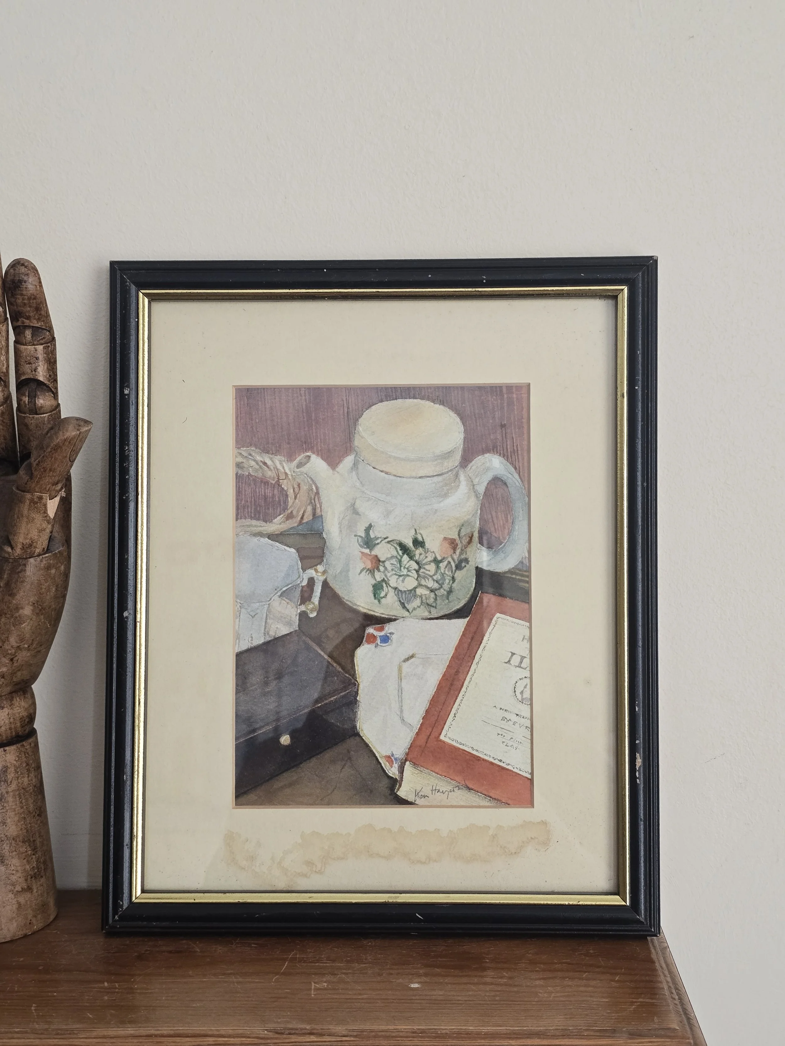 Signed original still life watercolour depicting a floral jug, folded textile and book in soft muted colours, presented in a vintage frame with aged mount.