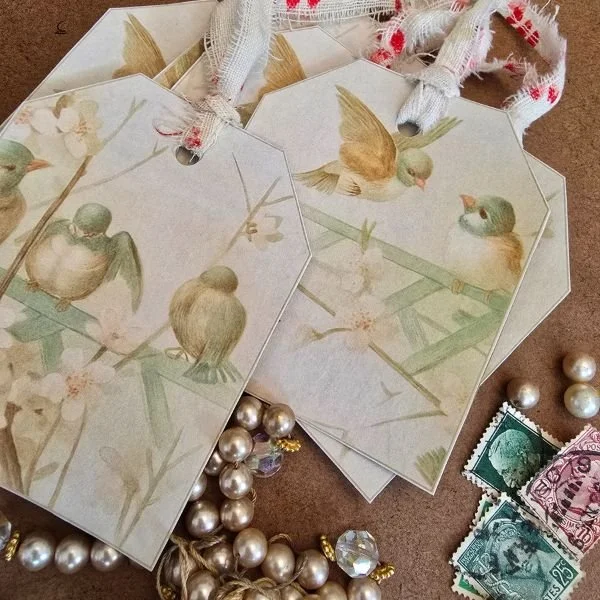 Set of handmade gift tags featuring delicate vintage-style bird illustrations on pale backgrounds, tied with frayed fabric ribbon with red detailing, styled alongside pearls and vintage postage stamps.