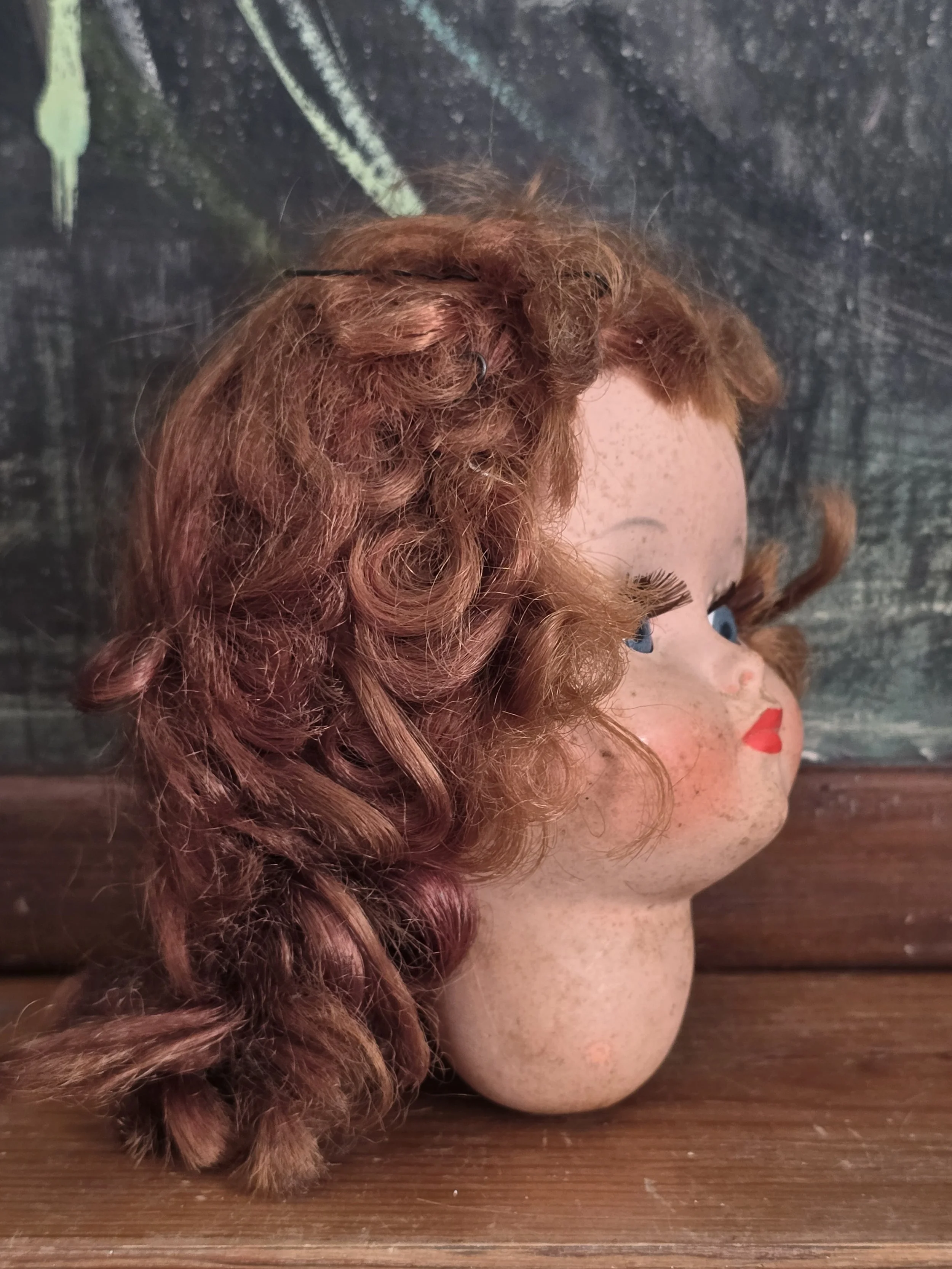 Side view of vintage French doll head with auburn curls and painted red lips, styled against a dark chalkboard background