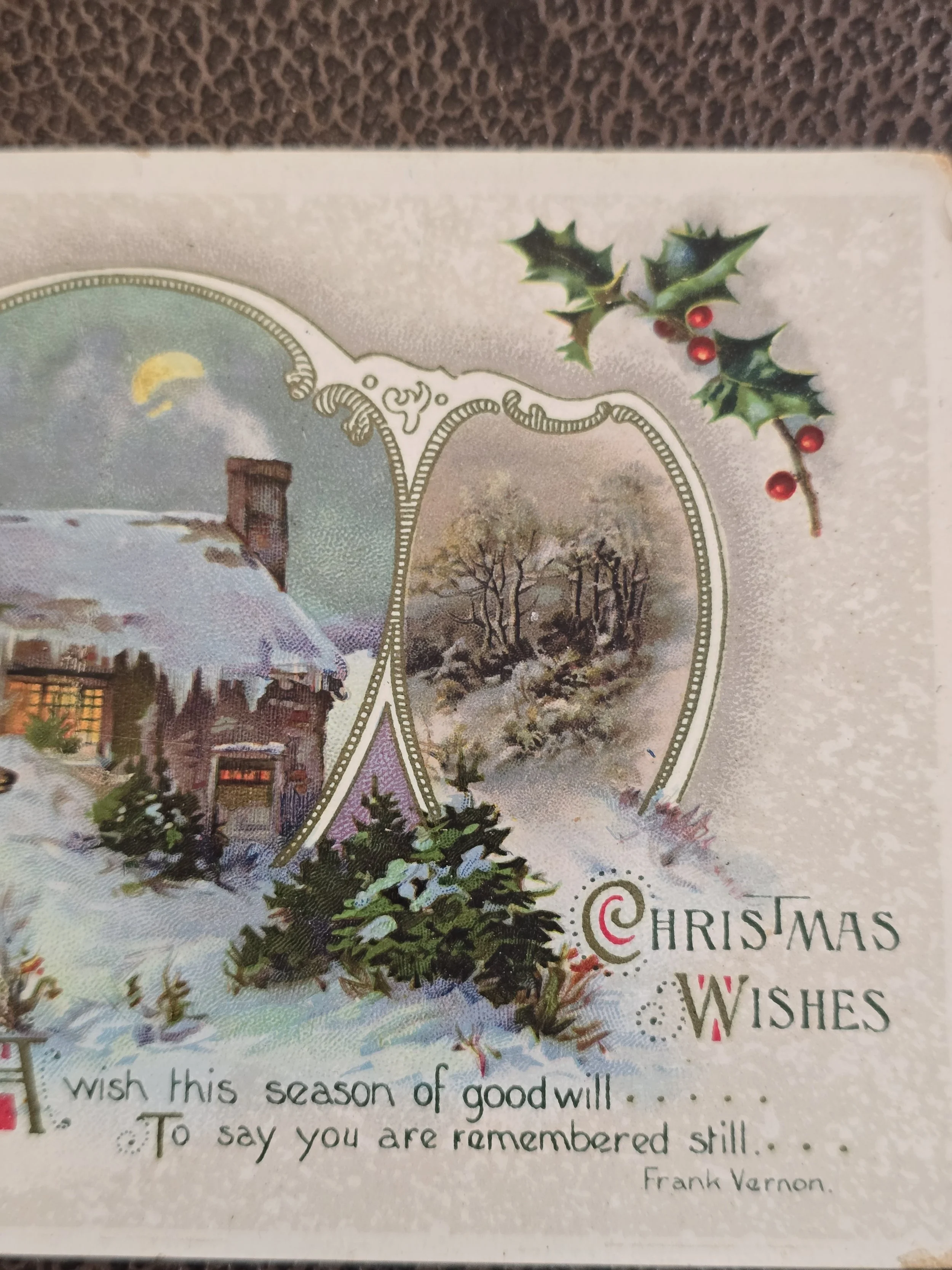 Close-up of antique Christmas postcard illustration showing a snow-covered cottage, holly berries and Christmas wishes verse by Frank Vernon.