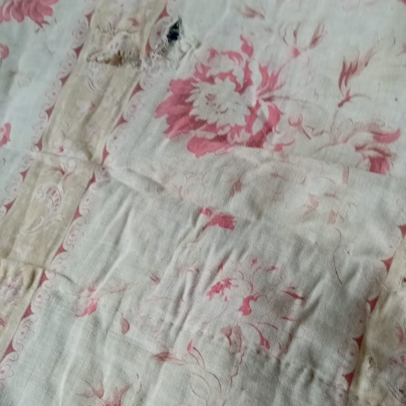 Close-up of a faded vintage floral textile in soft cream and pink tones, showing worn fabric, gathered stitching and age-softened colour. The delicate floral pattern and timeworn surface evoke memory, patina and the beauty of well-loved cloth.