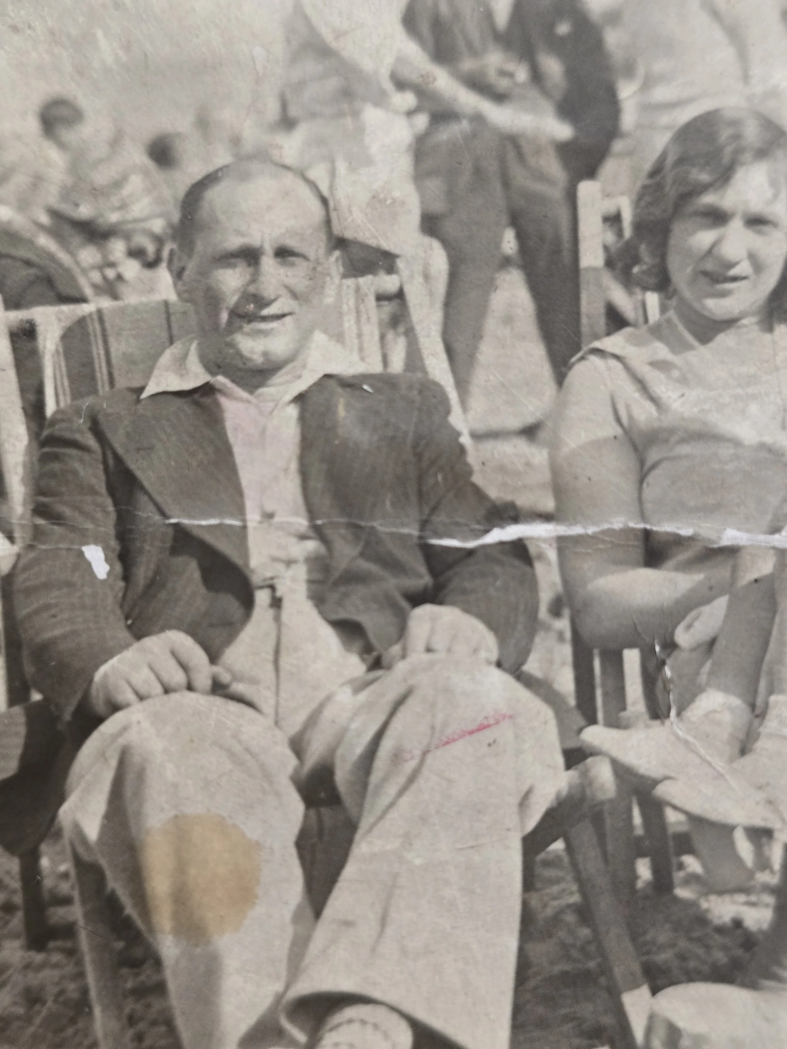 Timeworn vintage seaside photograph showing a family in deckchairs with holidaymakers on the beach behind them.