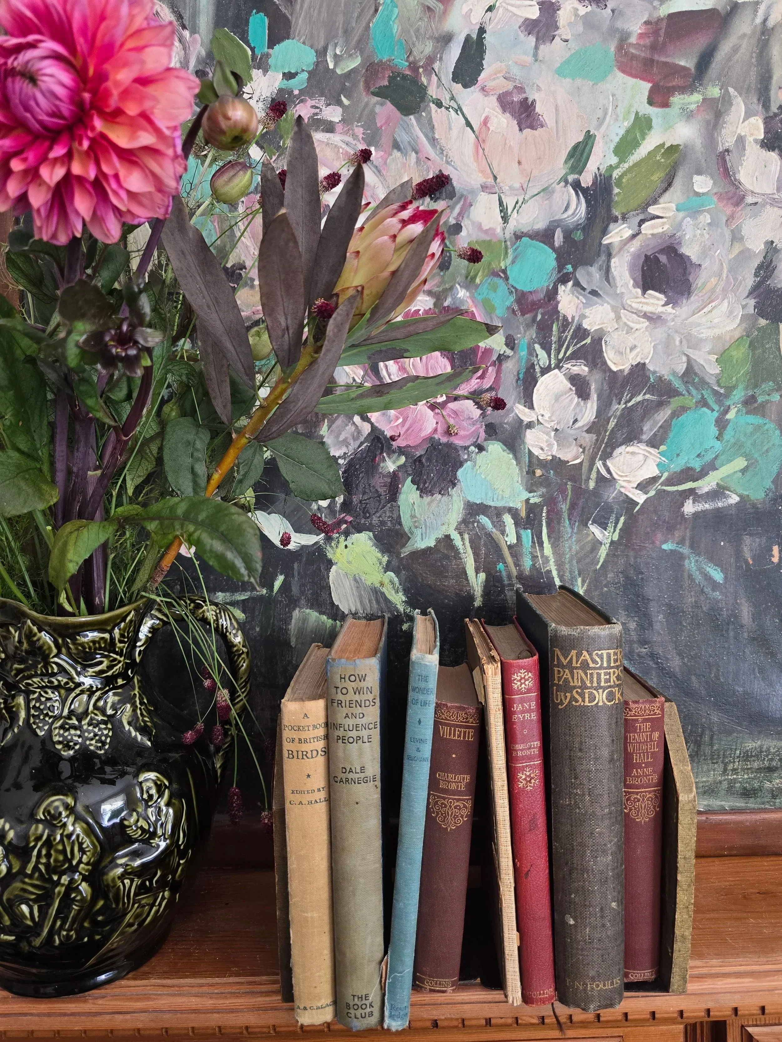 Vintage floral leather bookends holding a selection of old hardback books, including Charlotte and Anne Brontë titles, with floral painting in background.