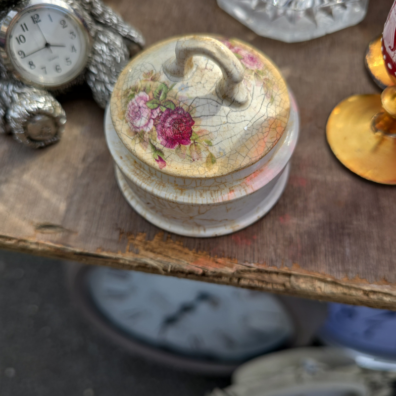 a small floral trinket pot with a crackled surface sits on a worn wooden surface. The lid is decorated with pink and burgundy roses softened by age.