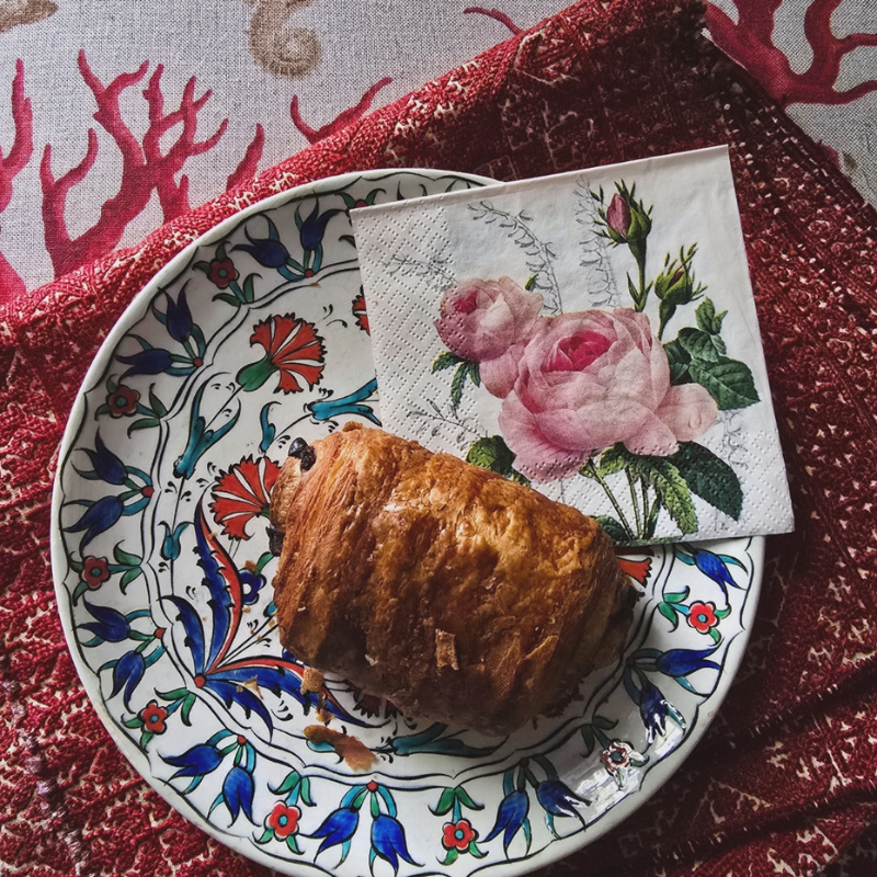 Pastry served on a colourful floral ceramic plate with a pink rose napkin resting beside it. The plate sits on a richly patterned red textile, creating a scene of everyday beauty, comfort and quiet domestic ritual.