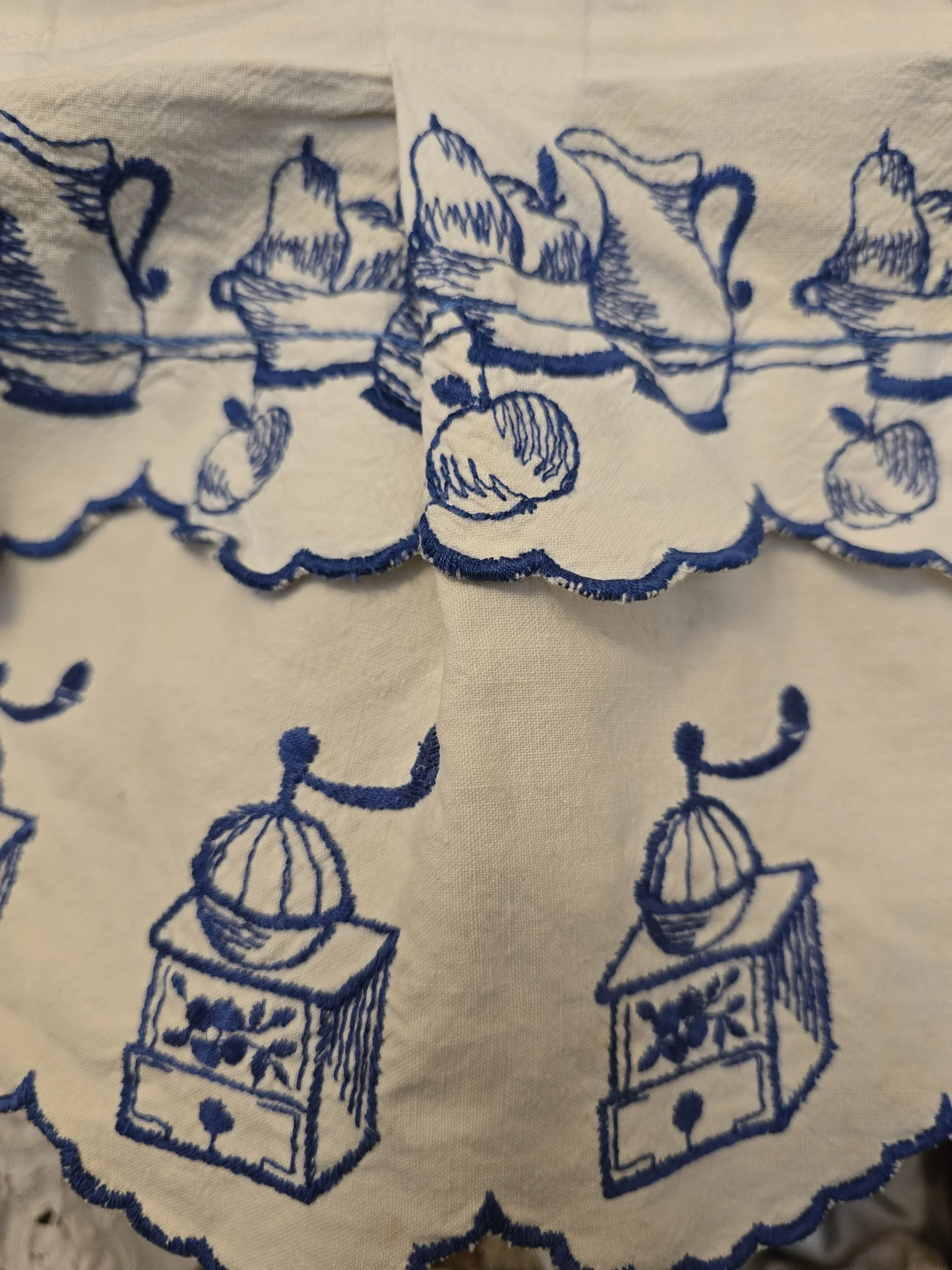 Detailed view of cobalt blue embroidery depicting jugs and baskets of fruit on an old white cotton kitchen pelmet.