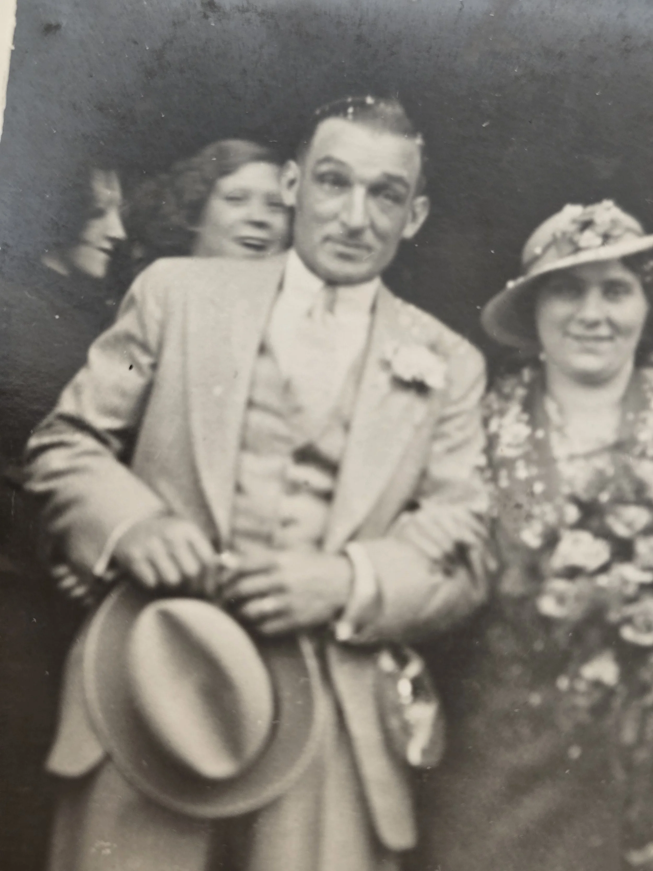 Close-up of a 1930s man in a pale three-piece suit holding his hat beside a woman carrying flowers.