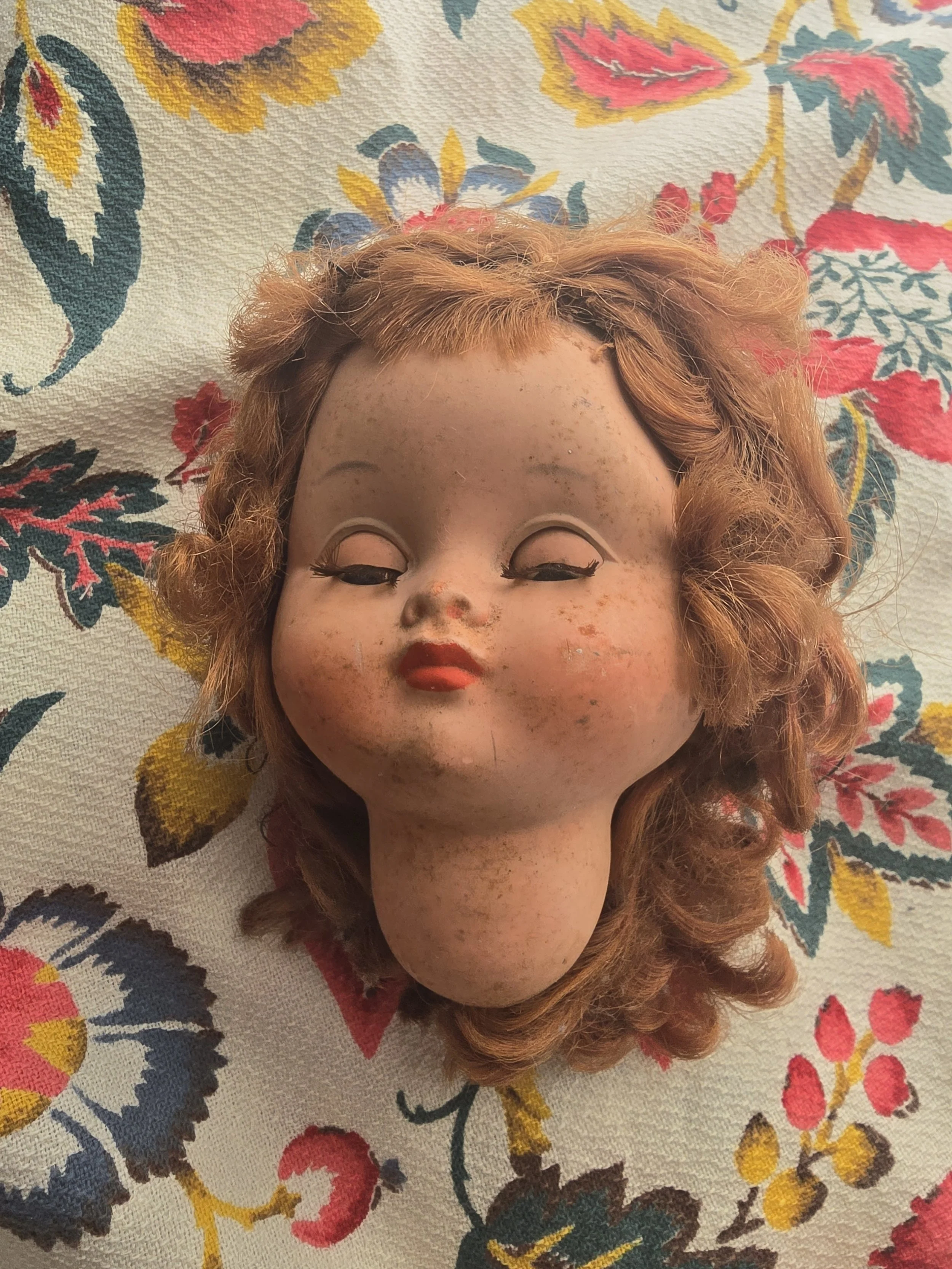 Vintage doll head resting on floral vintage fabric, showing curls and softly closed eyes in warm natural light