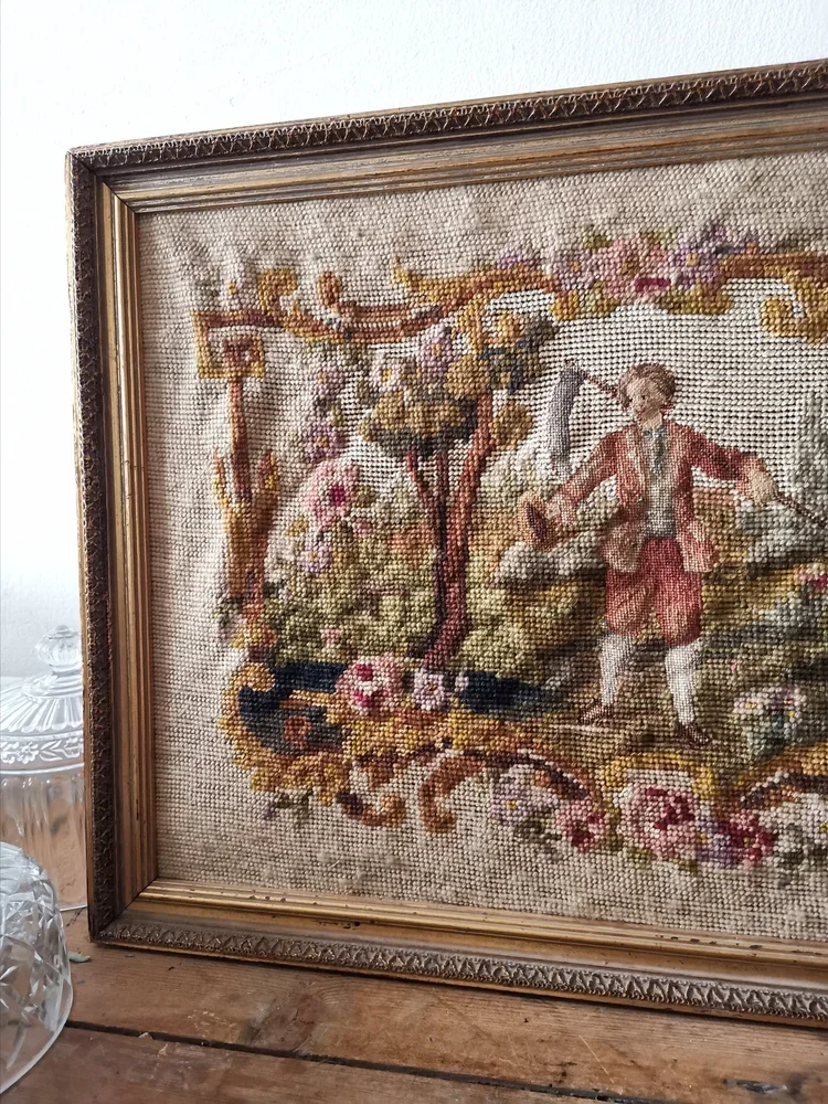 Framed needlepoint resting on rustic wood surface with vintage glassware