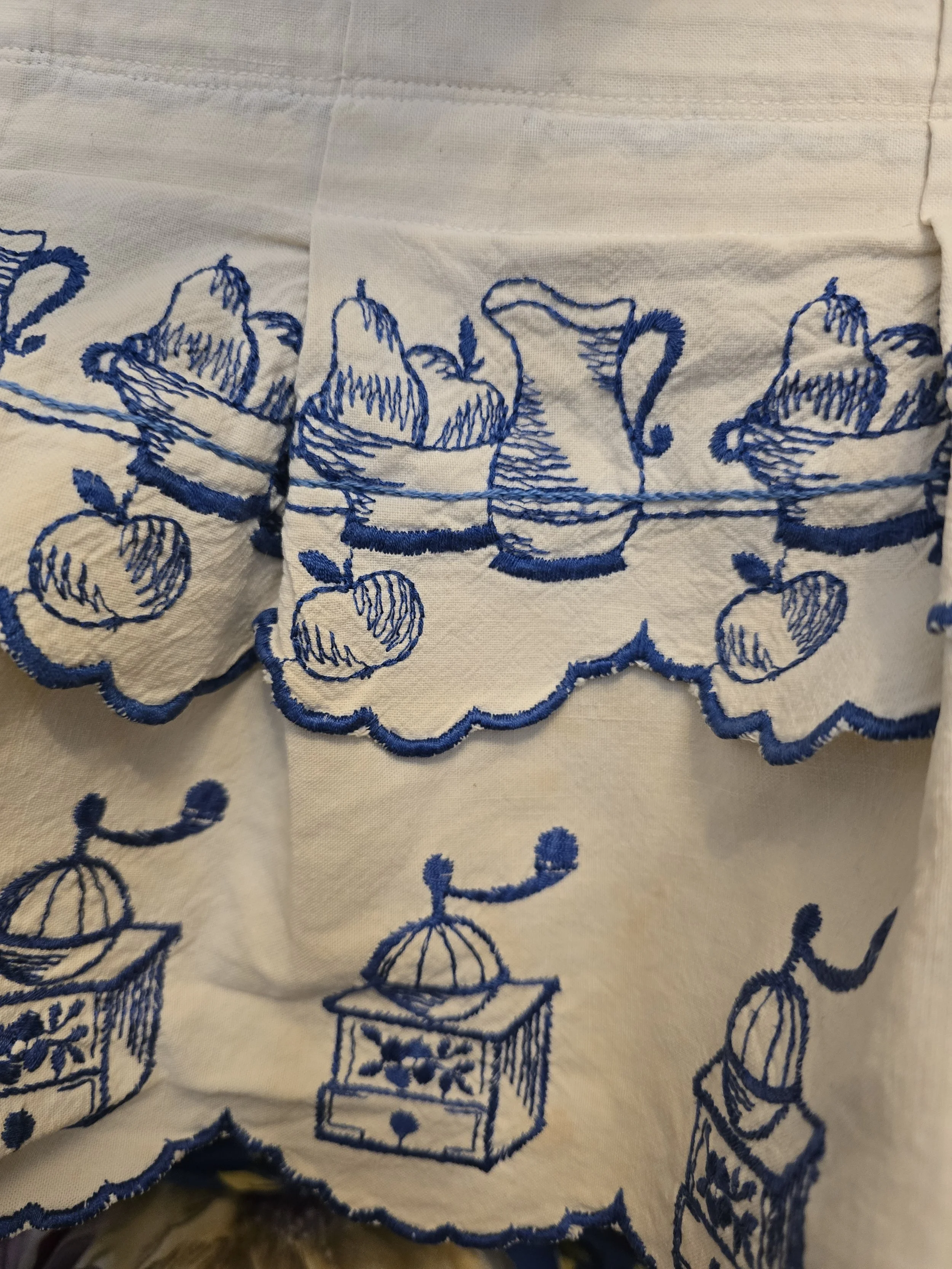 Blue embroidered vintage kitchen valance showing old-fashioned coffee grinders beneath decorative fruit and jug motifs.