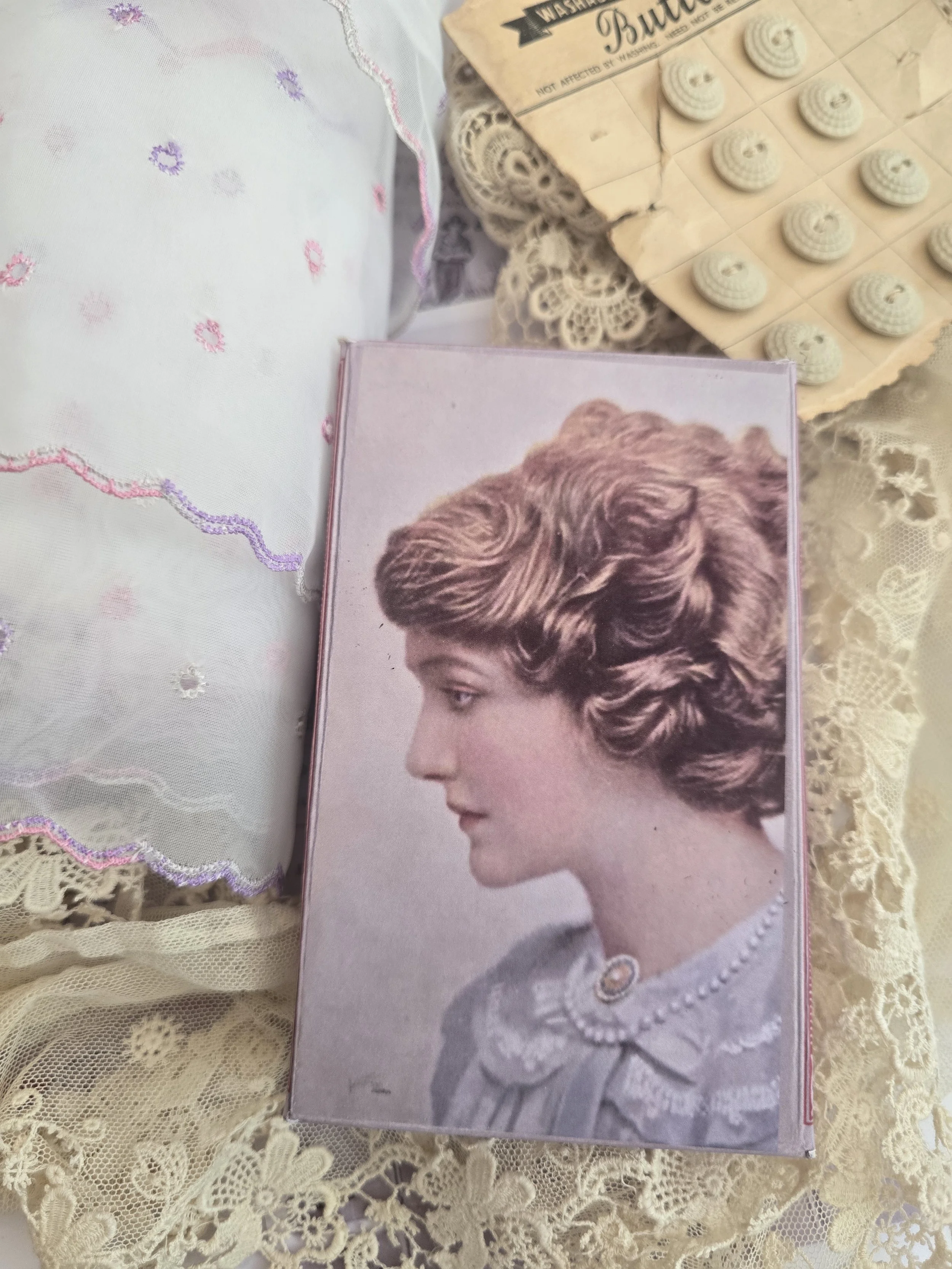 Handmade blush pink Pomme de Jour portrait box displayed amongst antique lace, embroidered textiles and vintage buttons.