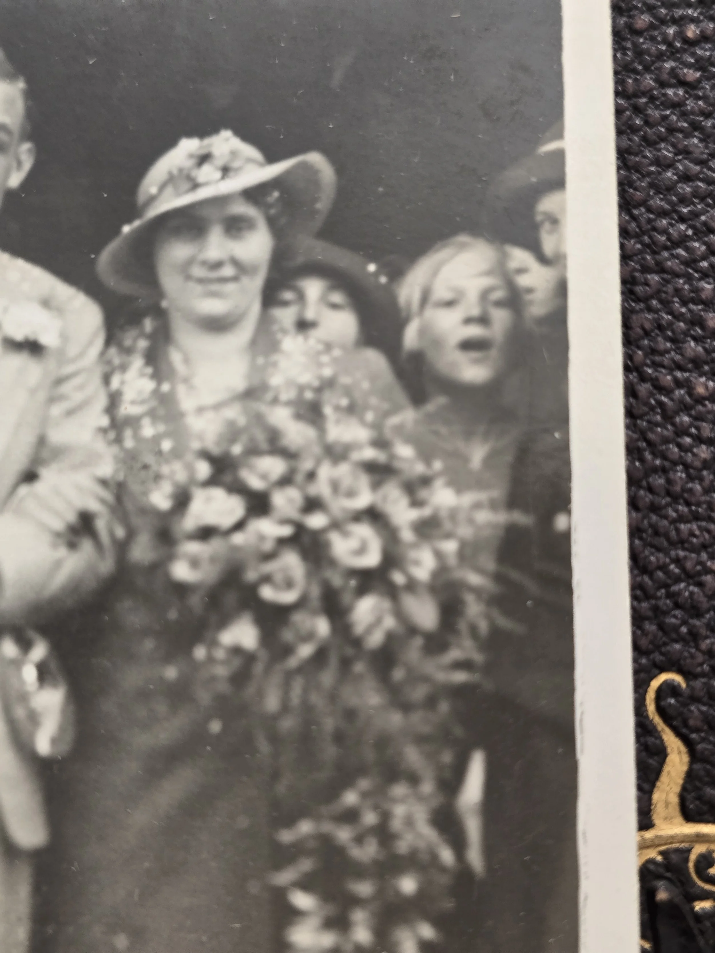 Detail of a 1930s woman in a flower-trimmed hat with a large cascading arrangement of flowers and people behind her.