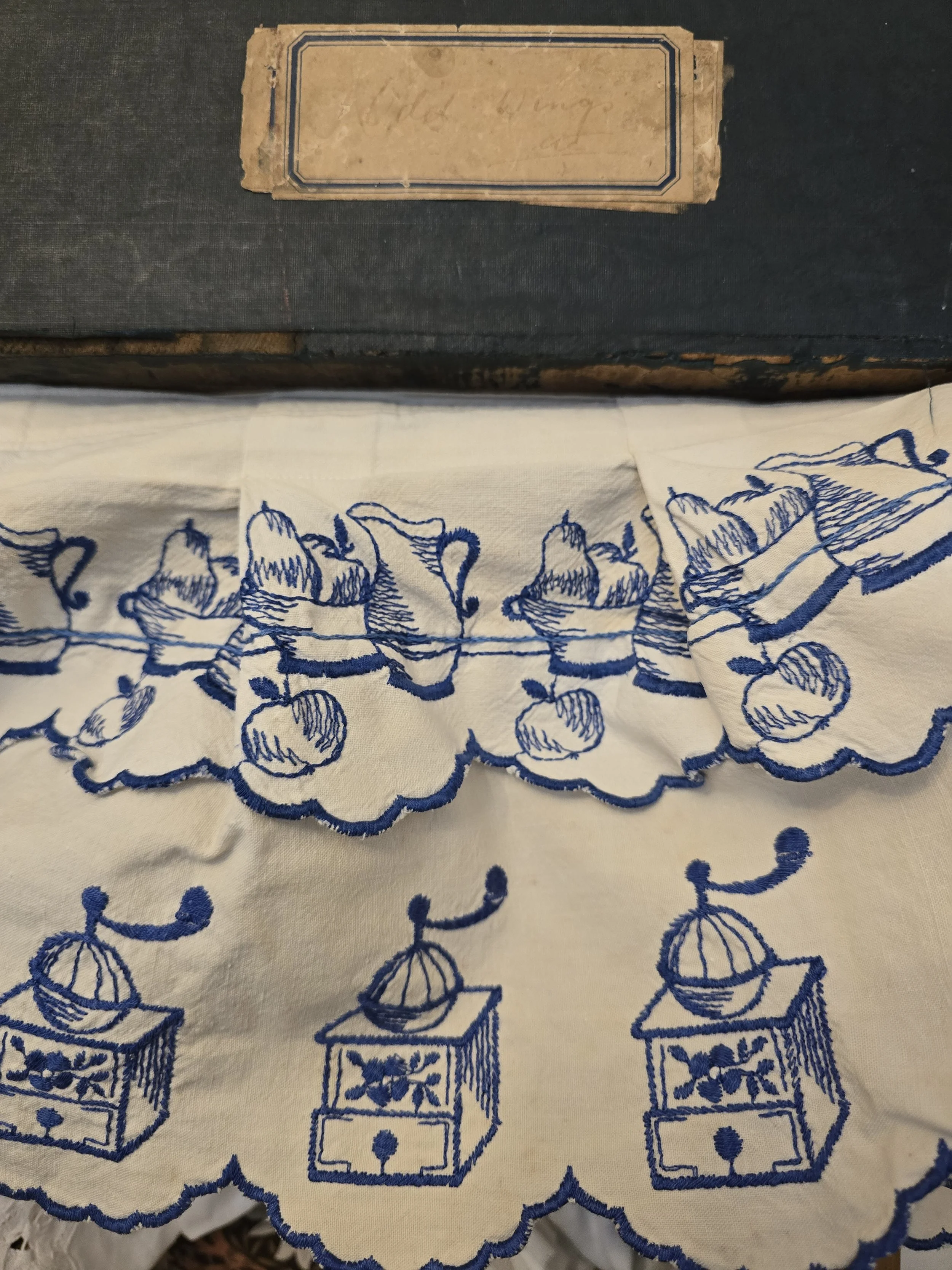Close-up of vintage white cotton kitchen valance with cobalt blue embroidery featuring a jug, fruit basket and scalloped edging.